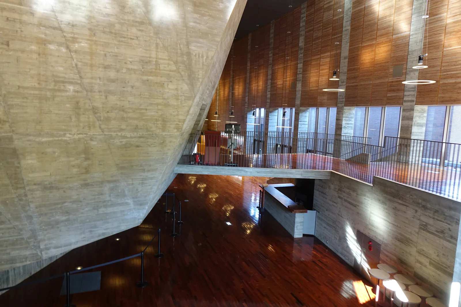自 foyer 上層俯視，左側是大片傾斜的清水混凝土壁面，右側是上層的走道與欄杆，下方是光亮的木地板與一座吧台，遠方是木板覆面的高牆與窗
