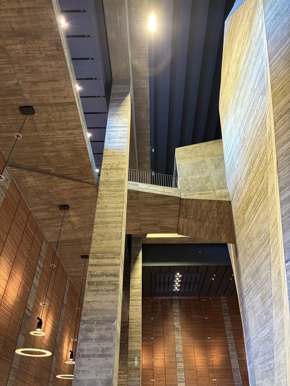 自 foyer 仰望，中央一根細長的混凝土柱從地面直達頂部，兩側是傾斜的混凝土壁面與木板牆，柱與柱之間架著上層的走道，頂部是深色的天花板與天窗