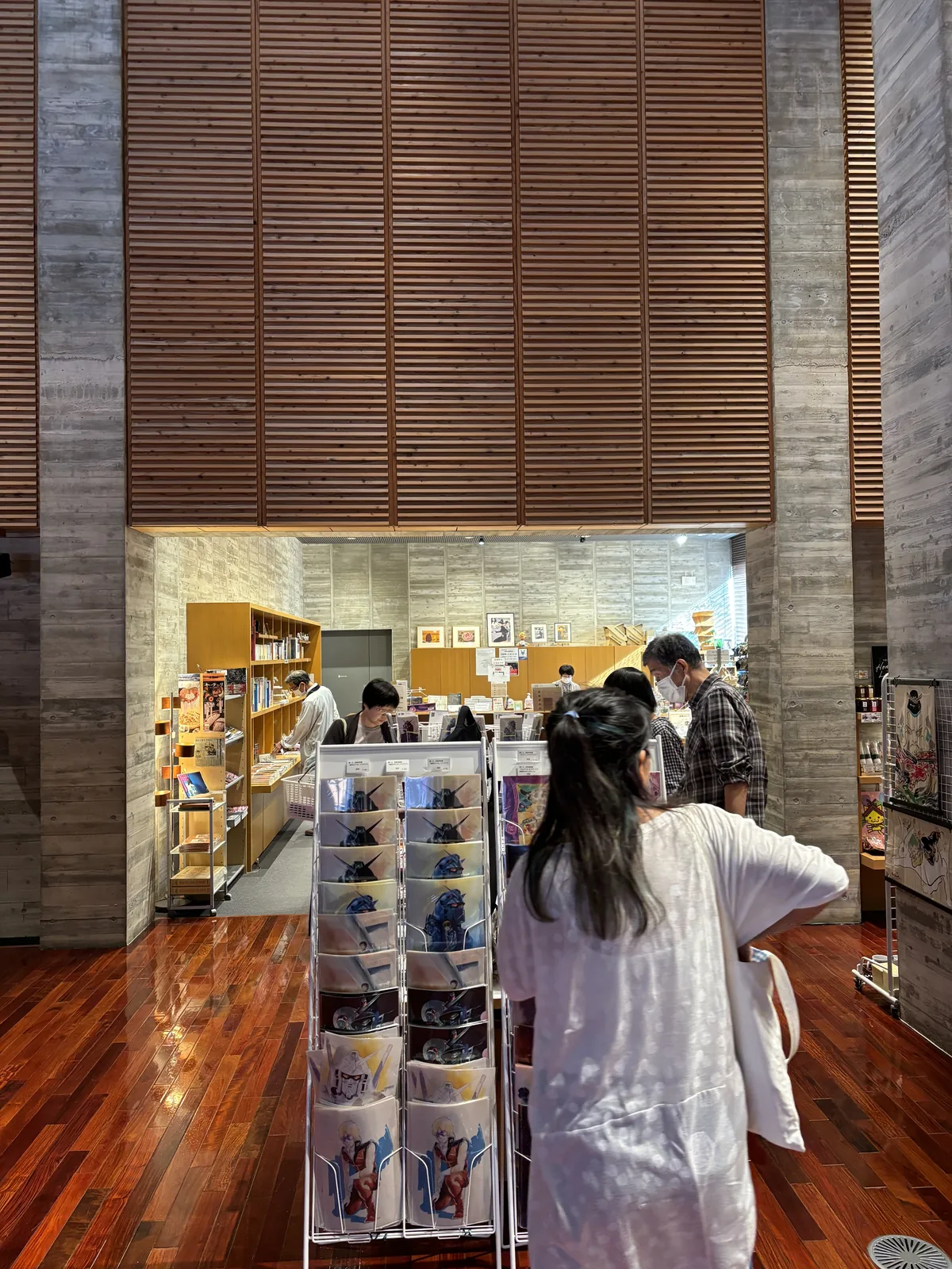 美術館大廳一角的 museum shop，上方是橫向木板的高牆與兩根混凝土柱，下方開口內是木製的商品架與參觀者，前景是一座擺滿明信片的旋轉架