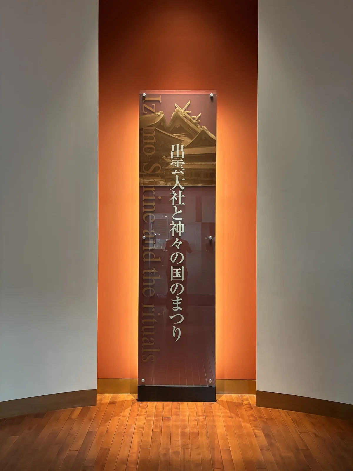 常設展示的入口，圓弧形的白色牆面圍出一個小前室，正面是一片橙色的牆，牆前立著寫有標題的透明板，地面是木地板，上下的間接照明打亮橙色的牆