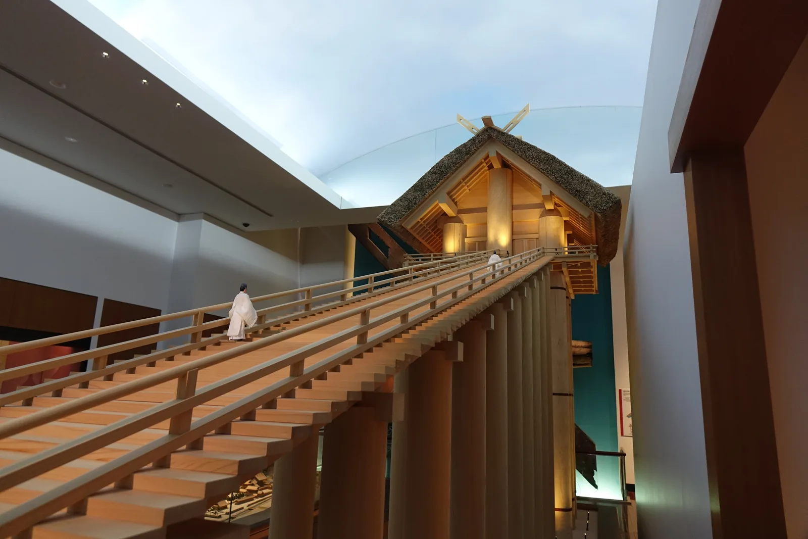 社殿模型的階梯近景，長長的木製階梯向右上方延伸到社殿，階梯下方是一排長柱，社殿的茅草屋頂在弧形的發光天花板下，左側可見另一處展示