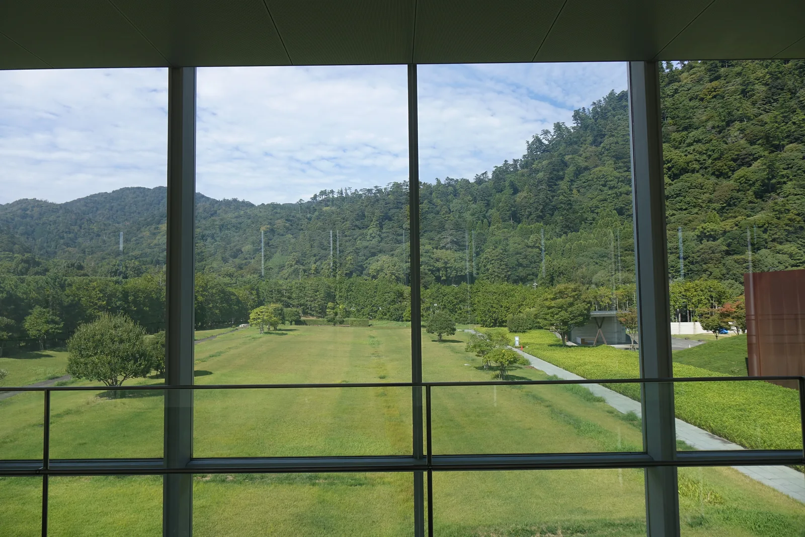 自上層隔著玻璃窗框望向庭園，下方是大片的草地與一條斜向的步道，零星的樹立在草地上，遠方是覆蓋森林的山，右側可見展示棟赤褐色的牆角