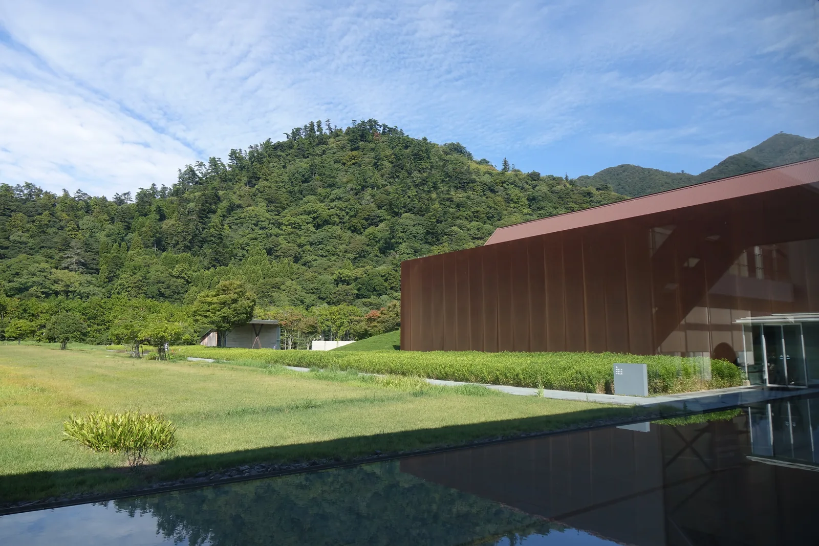 自大廳望向庭園，前景是一道淺水盤，水面映出天空，中景是草地，右側是一棟以赤褐色耐候性鋼板包覆的量體，背景是覆蓋森林的山與晴空