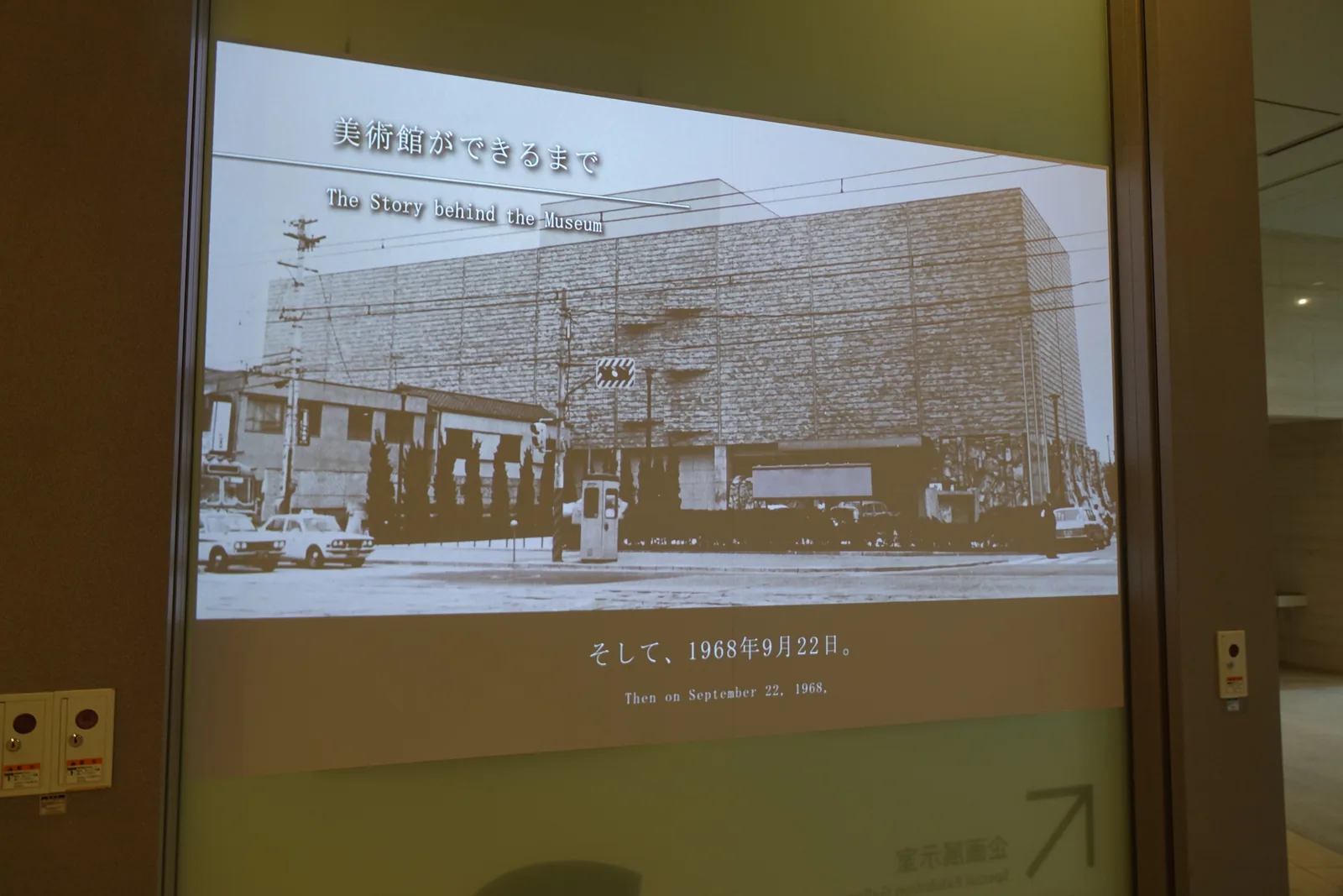 館內播放的介紹影像，畫面是一張黑白的舊館照片，一座以磚狀面材覆蓋、幾乎沒有開窗的方形建築立在街角，前方停著汽車，畫面上方與下方有標題與開館日期的文字