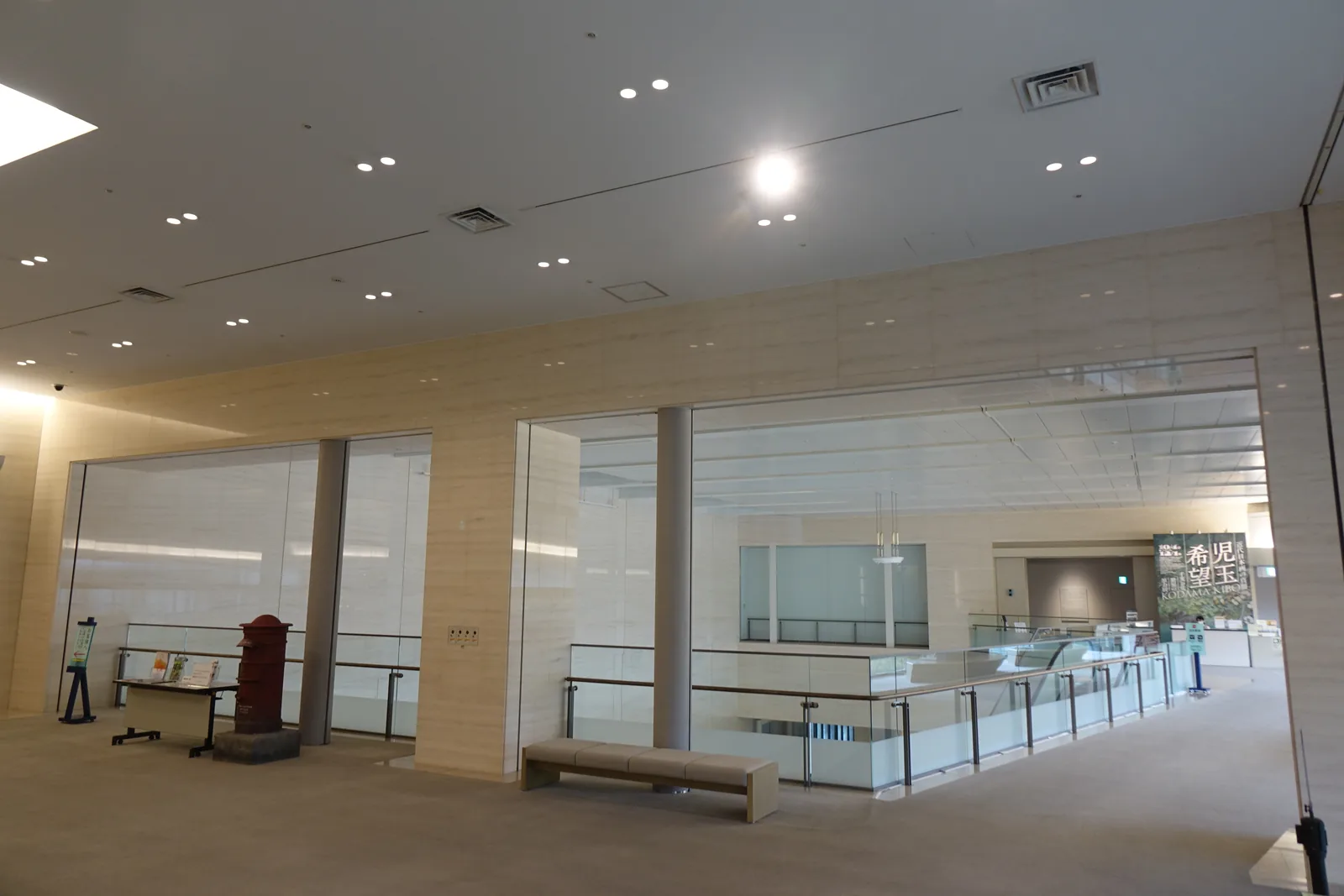二樓展示室前的空間，天花板以嵌燈均勻照明，牆面是淺色的面材，右側以玻璃欄杆向挑空開放，地面鋪地毯，中央一張長凳