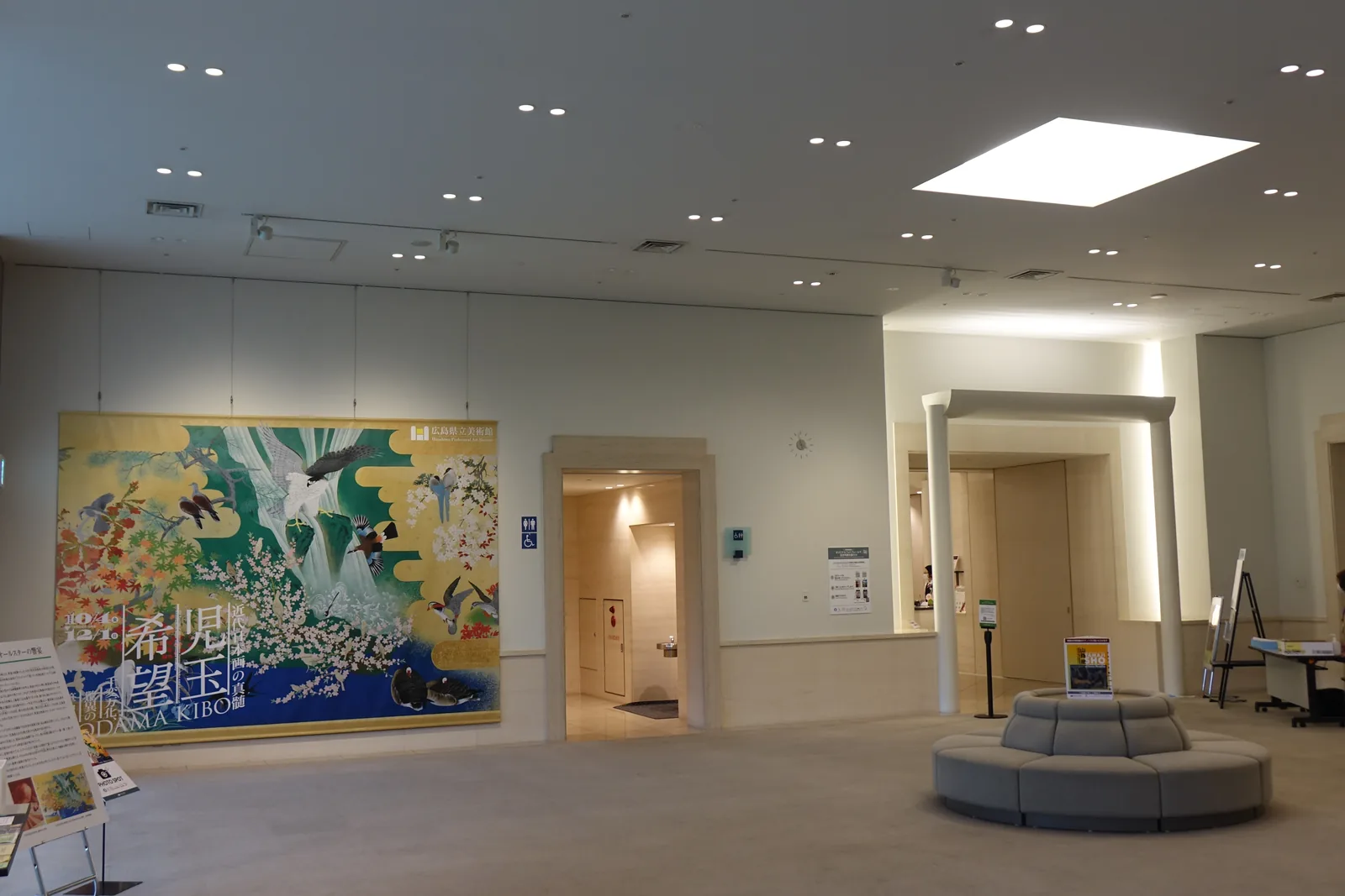 三樓企畫展示室的前室，天花板開著一片方形的天窗，正面是企畫展的大型看板與兩道以白色框收邊的入口，中央是一座圓形的長椅