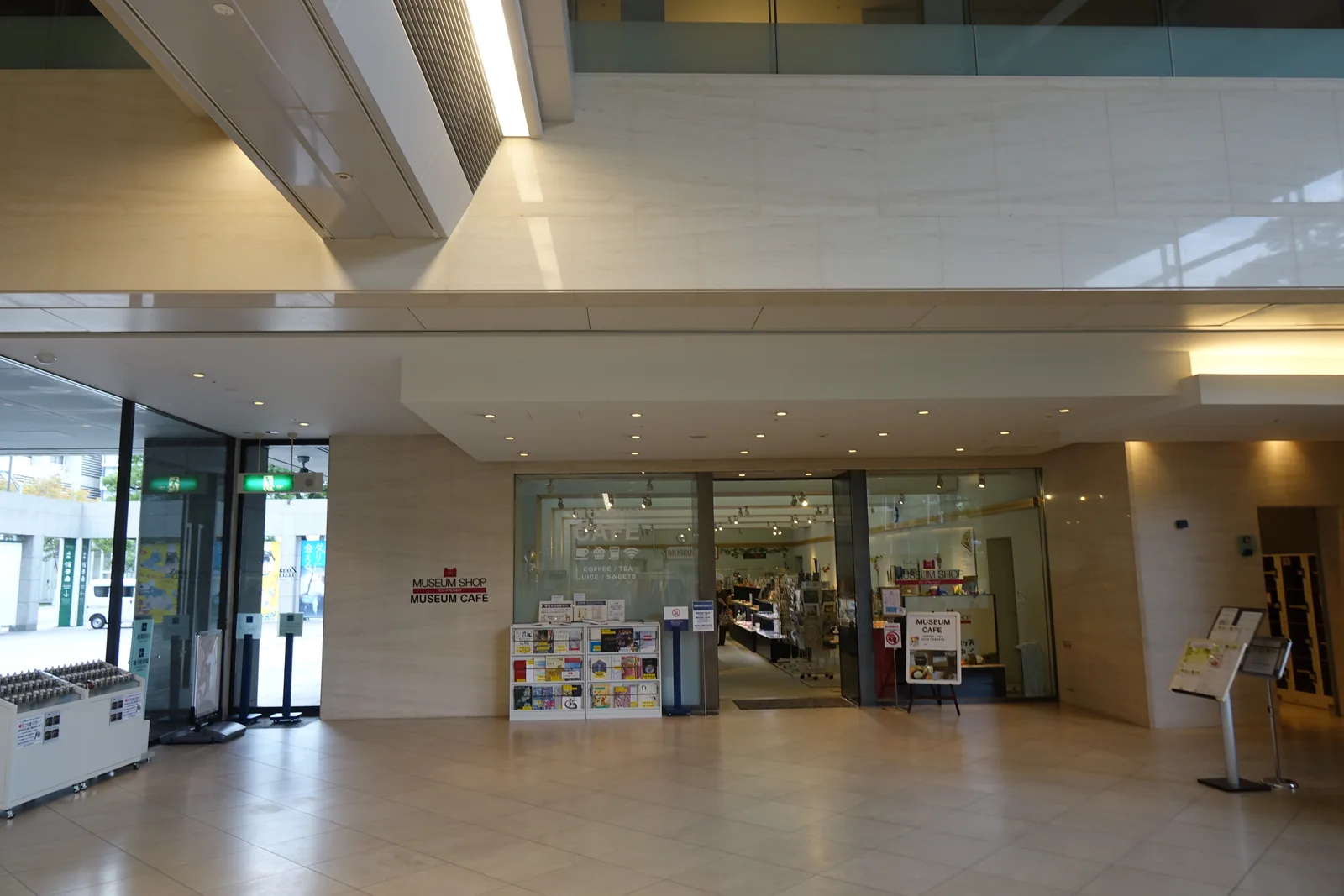 一樓的 museum shop 與 museum cafe 入口，以玻璃面向大廳開放，入口旁立著標示商店與咖啡館的看板與明信片架，上方是淺色的高牆與挑空的一角