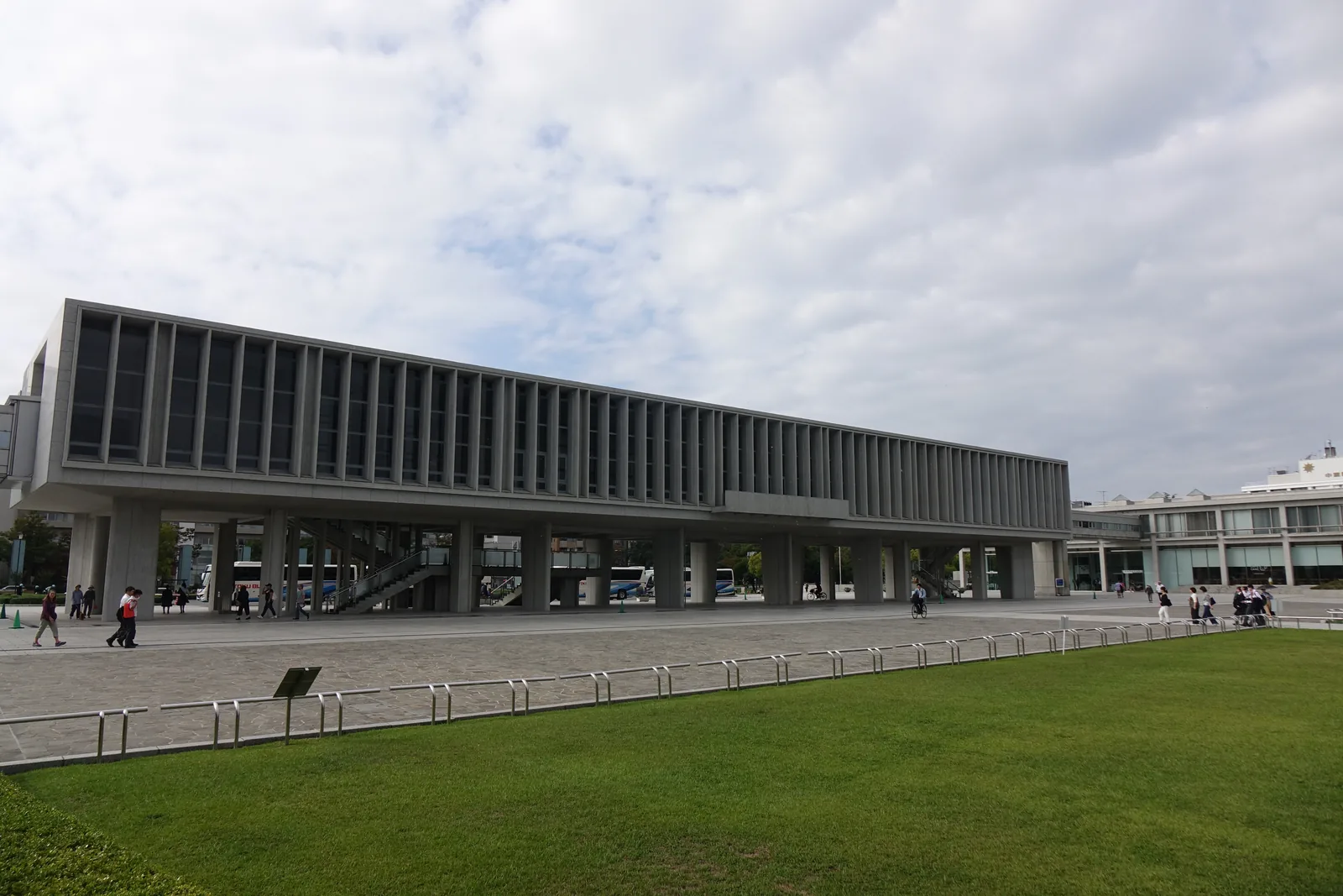 自北側草坪望向廣島和平紀念資料館本館，二樓是一條以竪向 louver 分割的長方形量體，一樓由一排柱子撐起而開放，柱間可見南側的街道與巴士，前景是草坪與低矮的欄杆