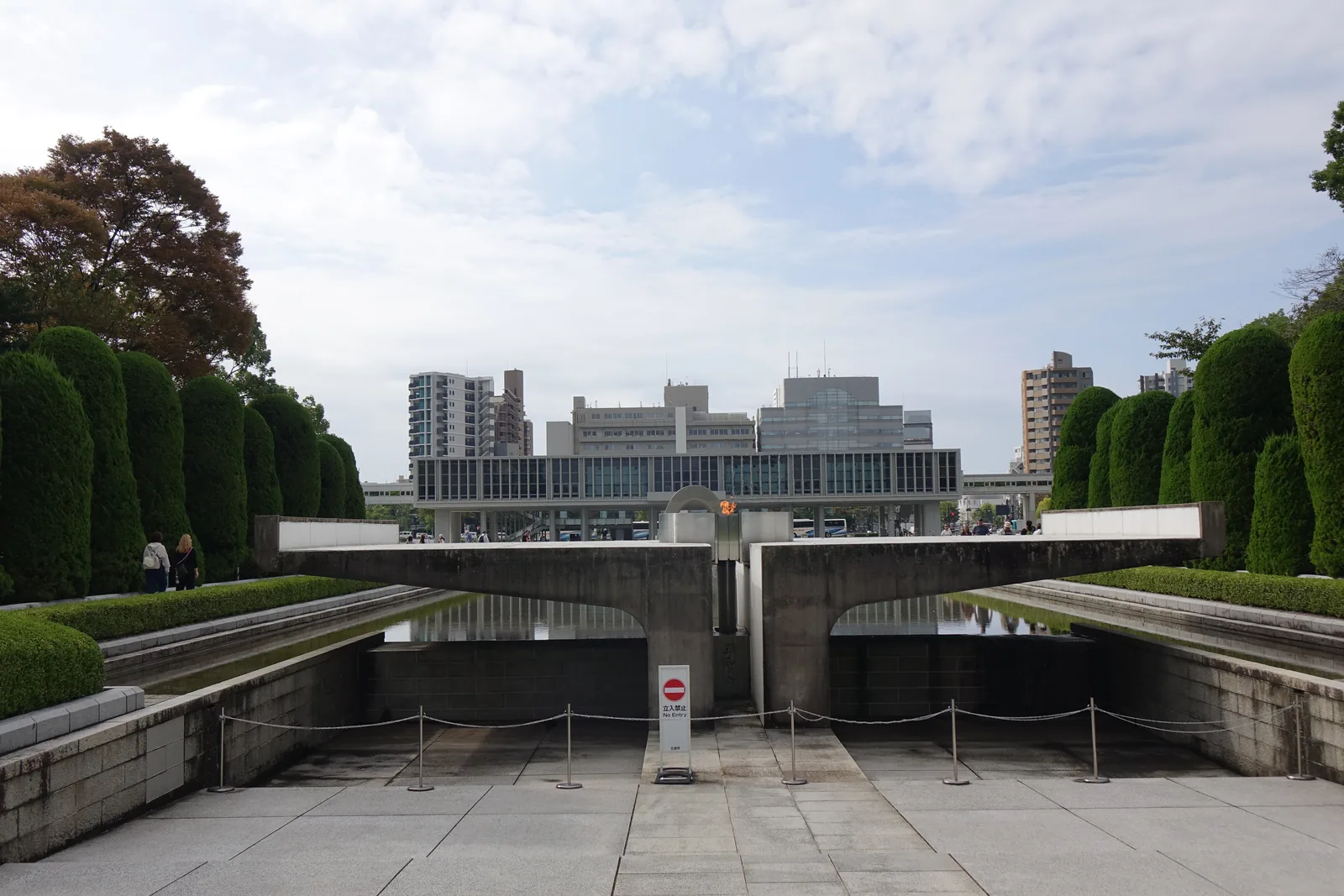 和平之燈與資料館本館，前景是以混凝土構成、向兩側張開的和平之燈與下方的水池，遠景是本館水平的量體與兩側的高樓，前方立著柵欄