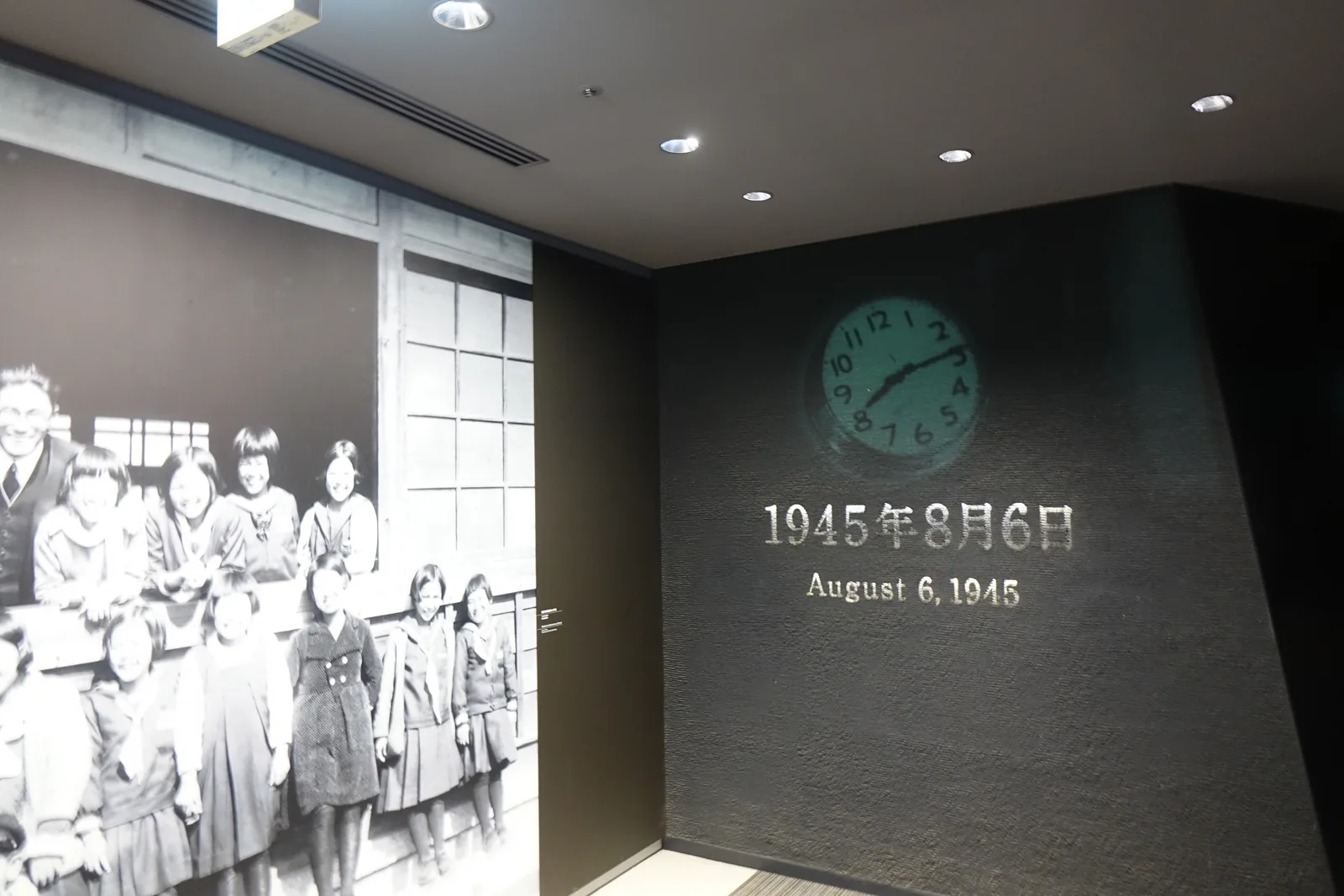 東館的導入展示入口，一面深色牆上嵌著一只綠色錶面的時鐘，下方寫著一九四五年八月六日的日期與英文，左側牆面是一幅放大的黑白團體照，天花板是深色的格柵