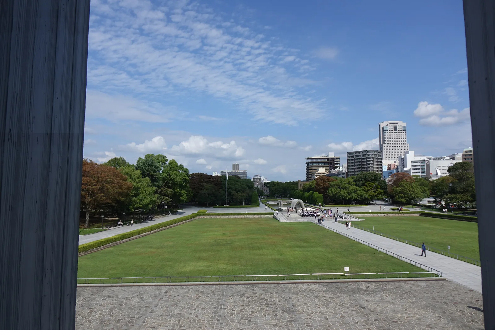 自館內隔著兩片粗厚的混凝土竪板向北望，窗外是石鋪的廣場、大片草坪與一條斜向的步道，中央是慰靈碑的拱與聚集的參觀者，遠處是原爆圓頂、旗桿與高樓，天空晴朗
