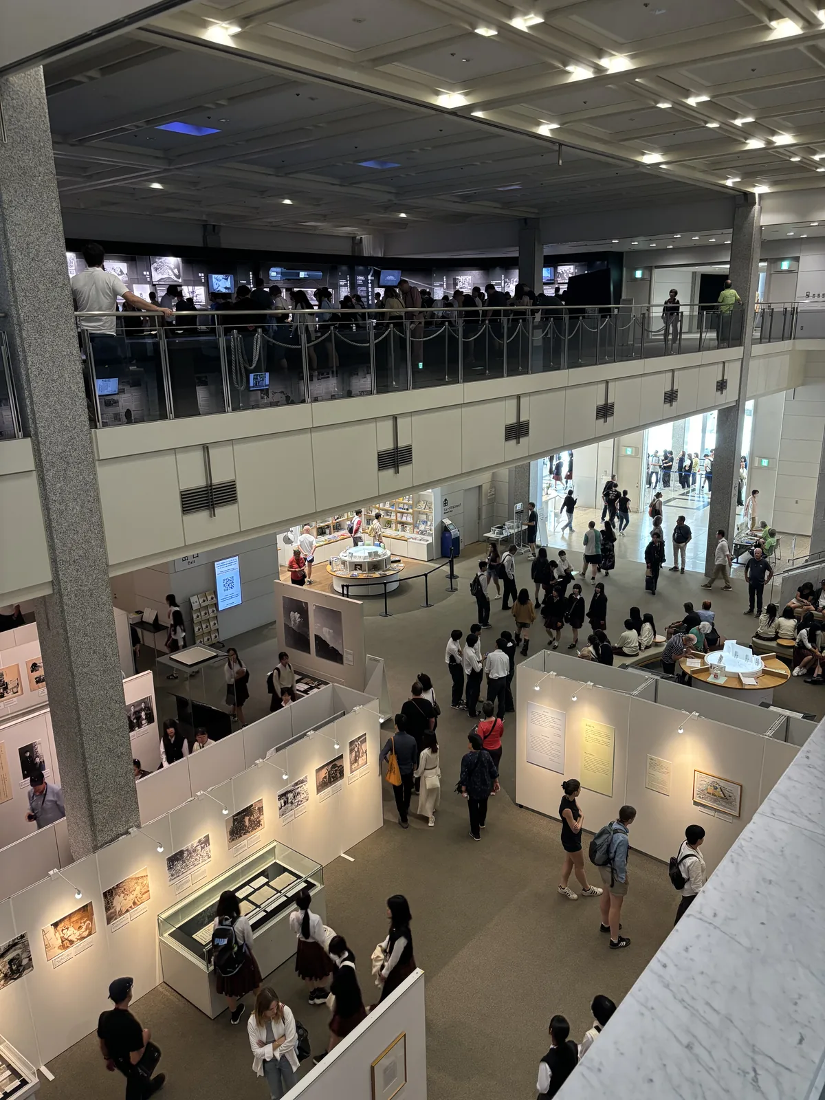 自東館上層俯視下層的展示區，畫面上方是另一側的迴廊與玻璃欄杆，下方是以白色隔板分隔的展示區、圓桌與大量參觀者，左側是一根石材包覆的柱子