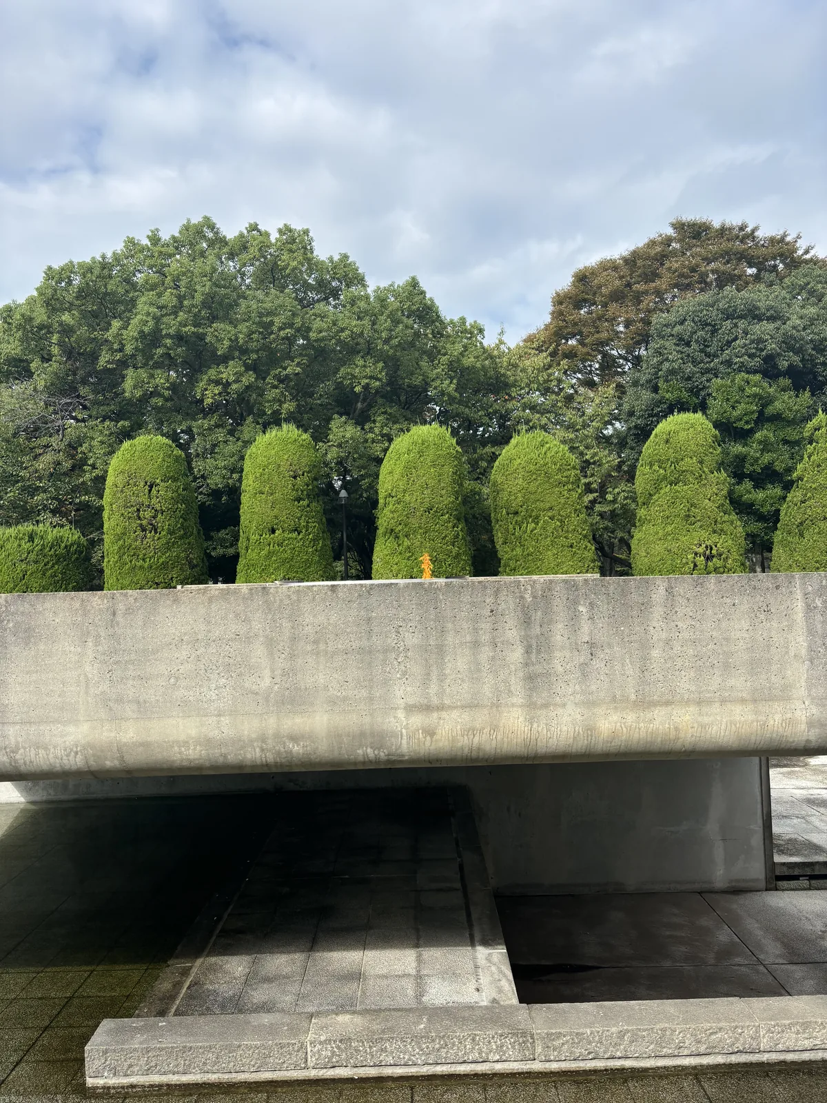和平之燈基座的近景，一道水平的混凝土量體以短柱架在水面上，表面留著模板的紋理，下方是陰影很深的開口，後方是修剪成圓柱形的樹籬與樹林