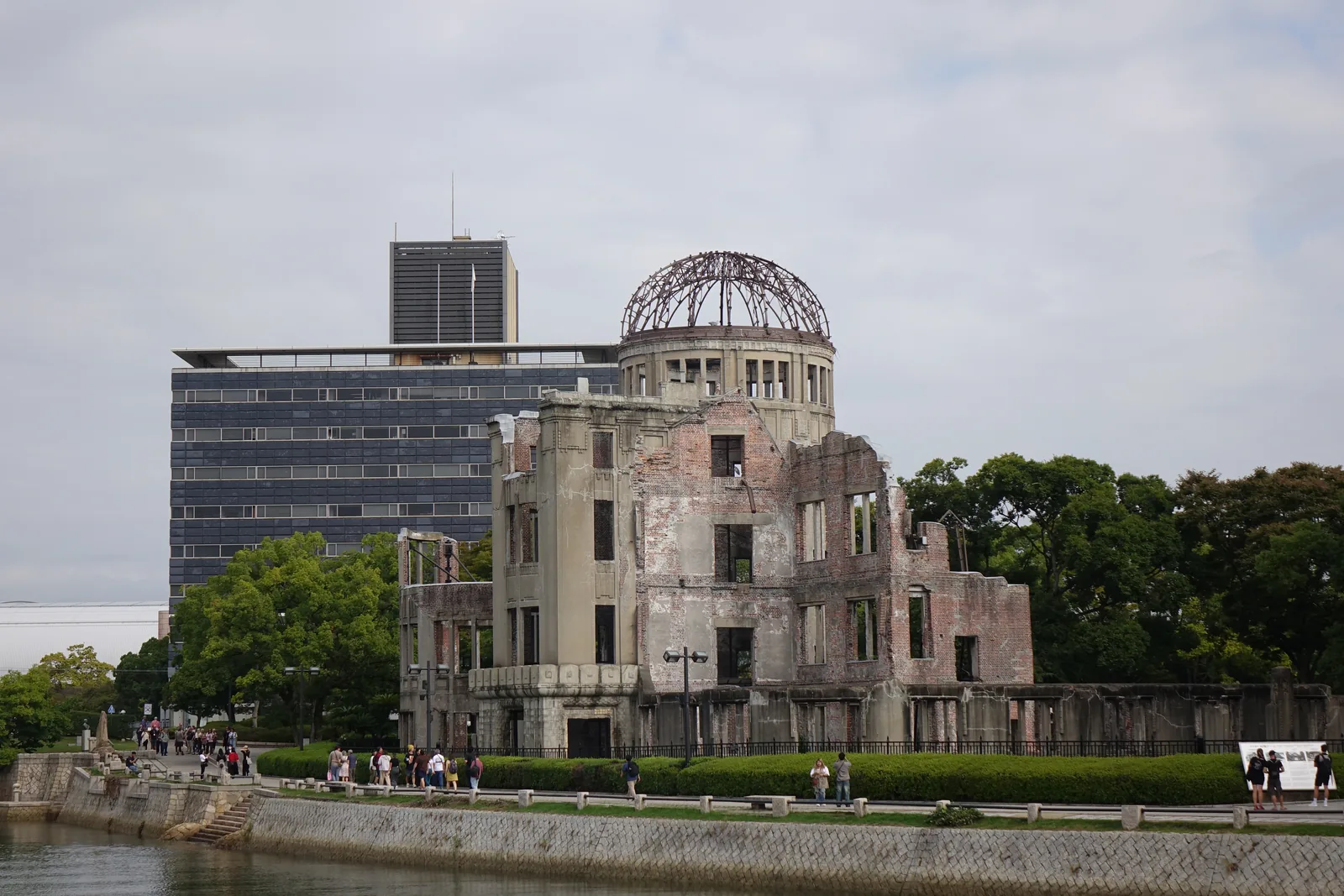 隔著元安川望向原爆圓頂，殘存的圓頂鋼骨架在一座剝落的磚造建築上，建築沿河岸展開，前景是平靜的河面，背後是一棟深色的高樓與另一棟橫向的辦公大樓