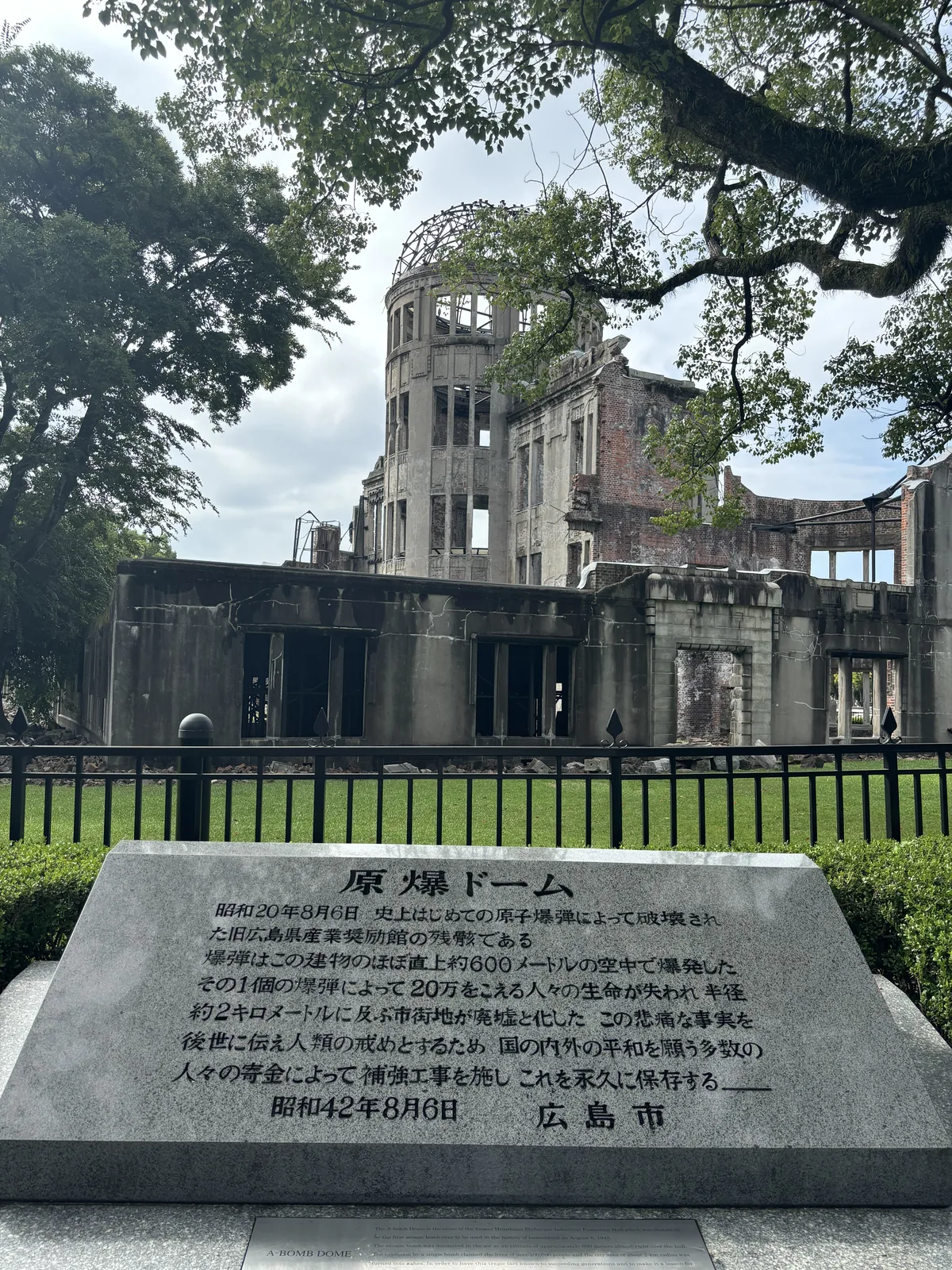 原爆圓頂北側的一塊石碑，碑上刻著關於原爆圓頂的說明文與昭和四十二年八月六日廣島市的落款，後方是柵欄內的原爆圓頂與一棵大樹的枝葉