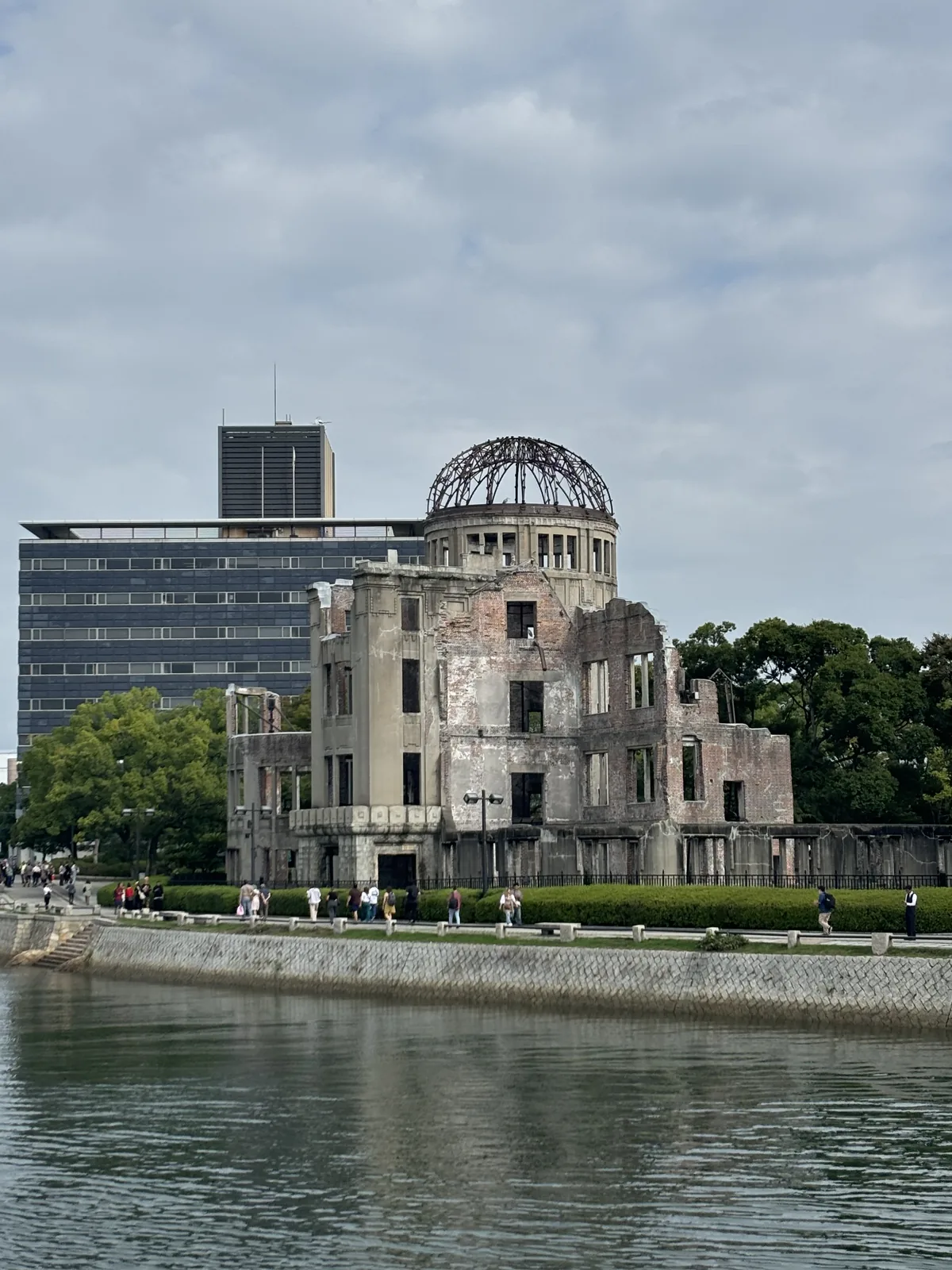 自元安川西岸的護岸望向對岸的原爆圓頂，圓頂與磚牆立在河邊的綠帶上，後方是一棟深色的高樓與一棟橫長的辦公大樓，前景是河面
