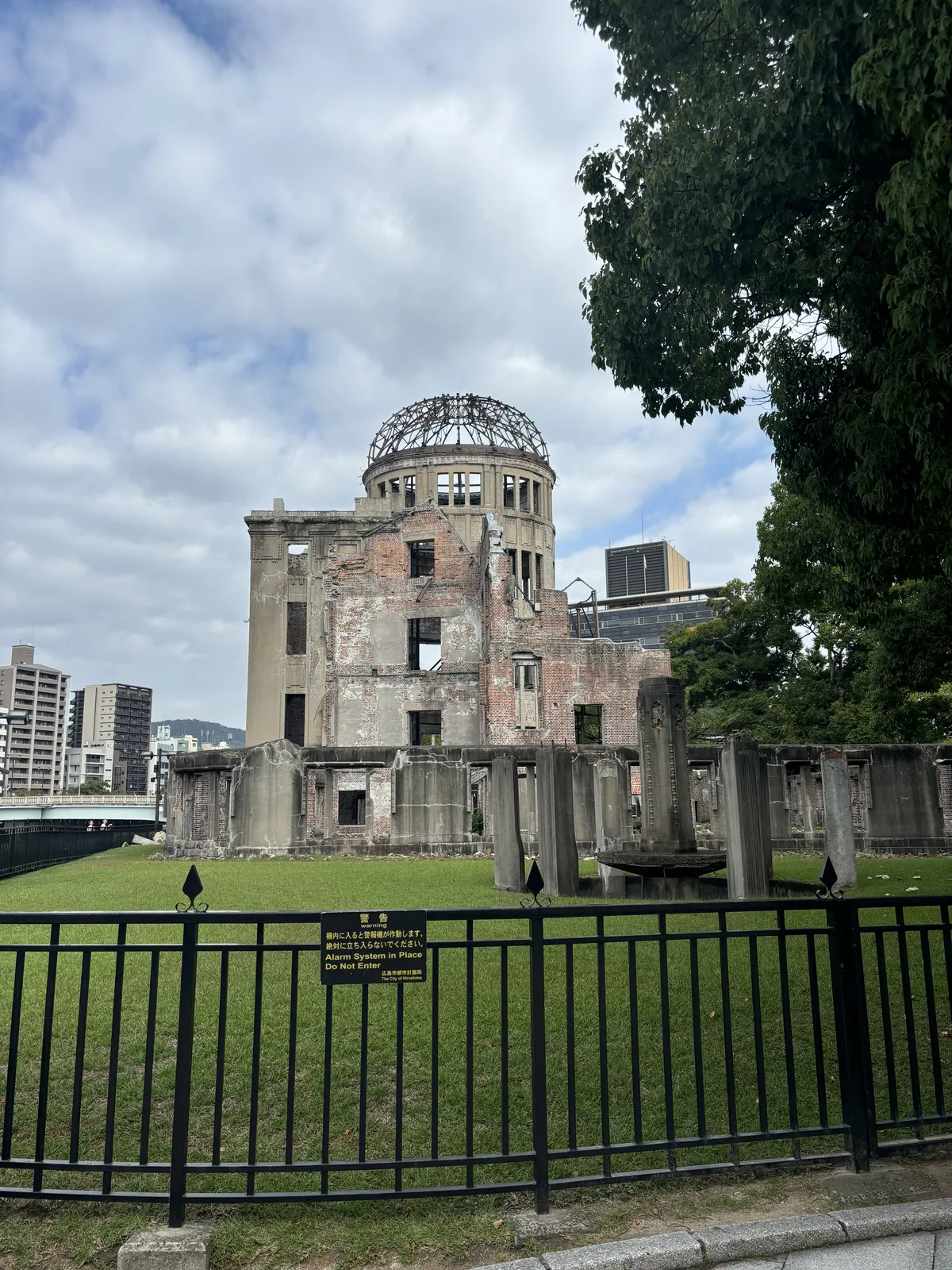 自北側隔著黑色鐵柵望向原爆圓頂，柵內是草地與散落的殘存牆柱，圓頂立在後方，右側是一棵大樹的枝葉，左遠方是高樓