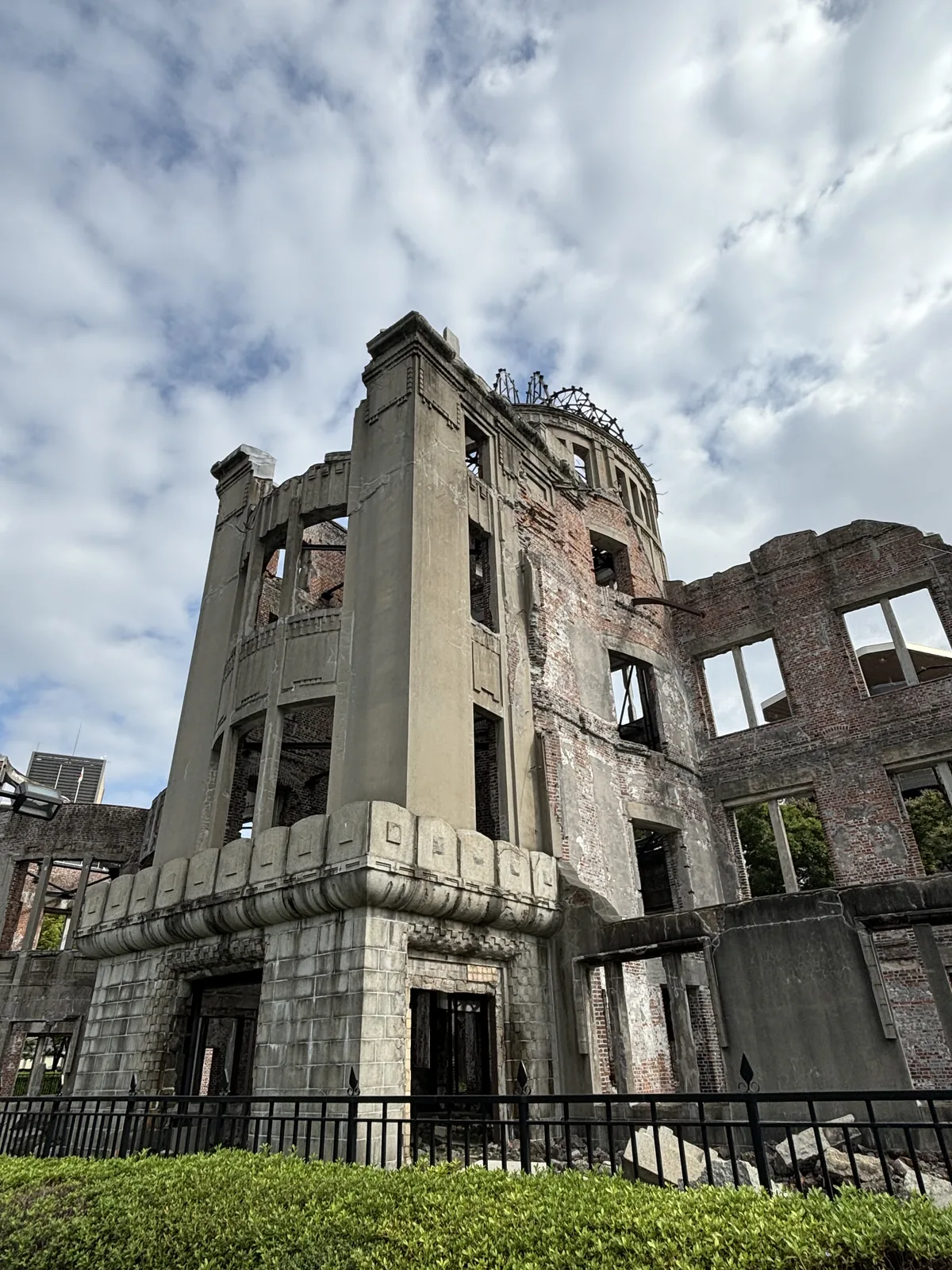 原爆圓頂轉角量體下段的近景，粗石砌成的基座與一道凸出的線腳，上方是磚牆與門窗的開口，表面殘留著灰白色的砂漿，前方是黑色的鐵柵