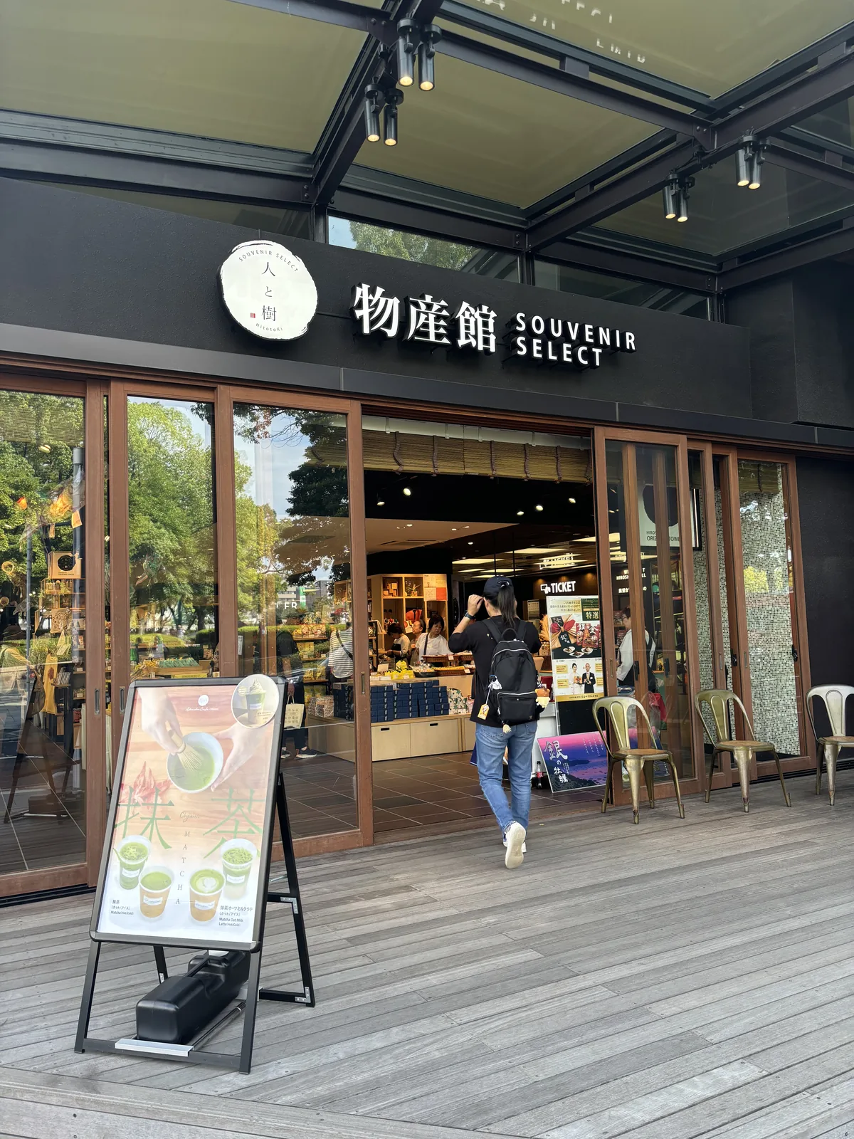 一座商店的入口，上方以白字寫著物產館與英文，入口前是木平台，一位參觀者正走進店內，店內可見陳列架與商品