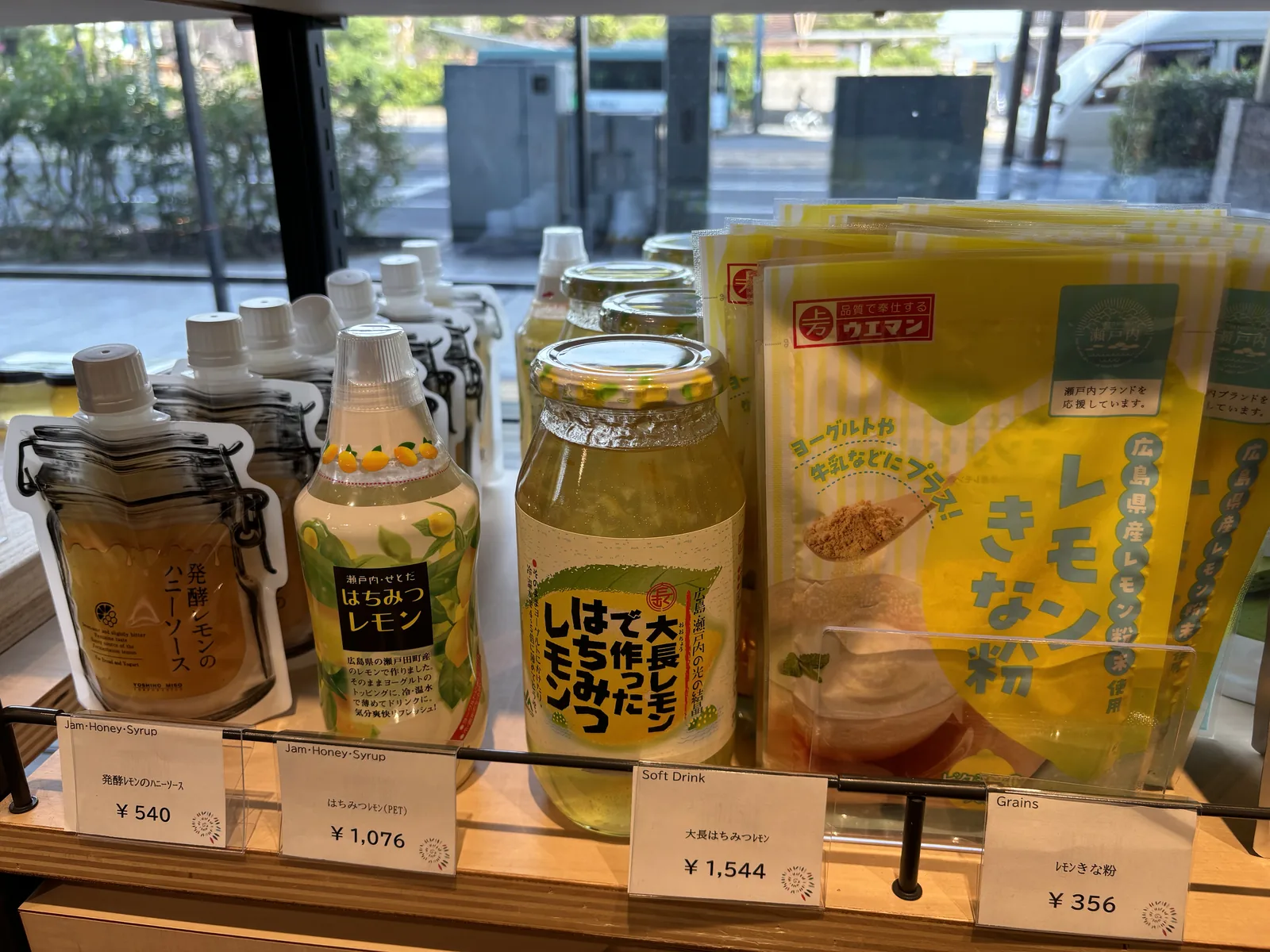 物產館的陳列架上擺著以廣島檸檬製成的果醬、蜂蜜檸檬飲料與檸檬黃豆粉，標價牌寫著價格，背後的玻璃外是街道與樹