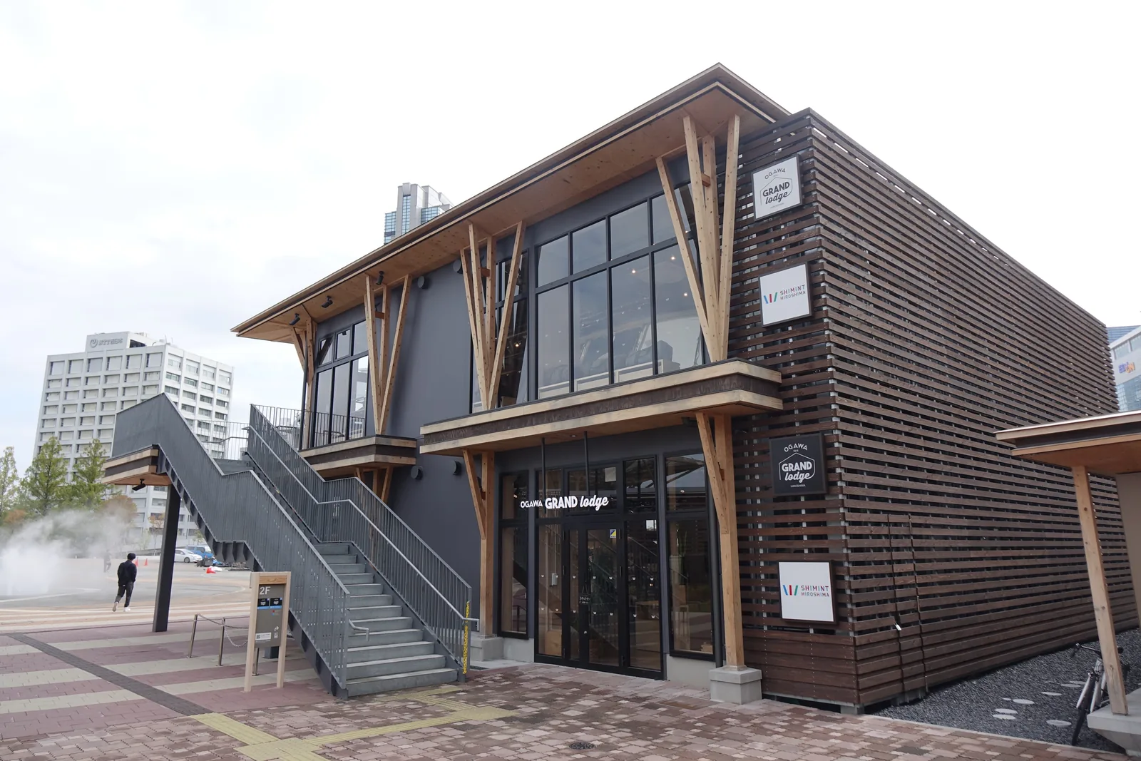 廣島 Gate Park 的一棟木造店舖，兩層的量體以深色的木百葉包覆，外周以分叉的樹狀木柱撐起深簷，側面有一道鋼製的外部樓梯，前方是磚紅色的鋪面