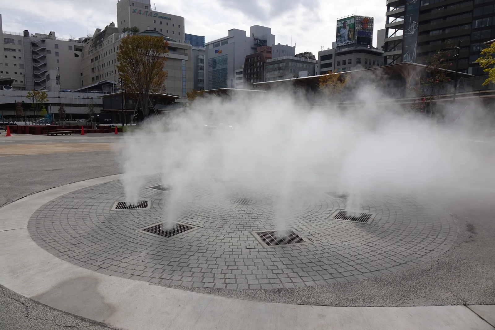 廣場上的噴霧噴水，圓形的石鋪地面上有數個噴口，白色的霧氣升起，背後是市區的高樓