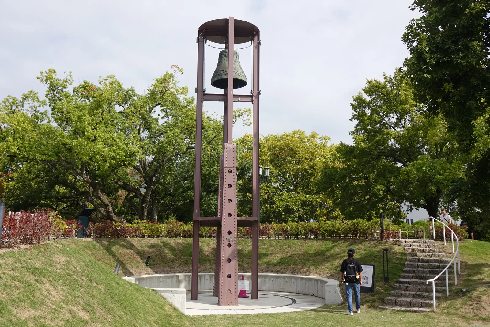 和平之鐘，一口青銅鐘懸在一座細長的鋼製鐘架頂端，鐘架下方是圓形的下沉廣場，後方是草坡、樓梯與樹林