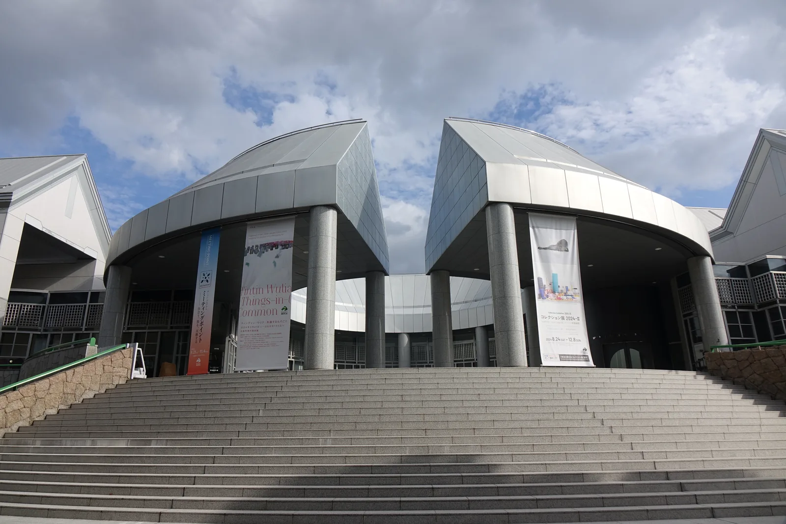 自石階下方仰望廣島市現代美術館的正面，寬大的石階通往一個以圓柱撐起的弧形屋簷，屋簷兩側各是一座以銀色金屬板覆蓋、山牆朝前的切妻屋頂，屋簷下懸著展覽的垂幕，天空多雲