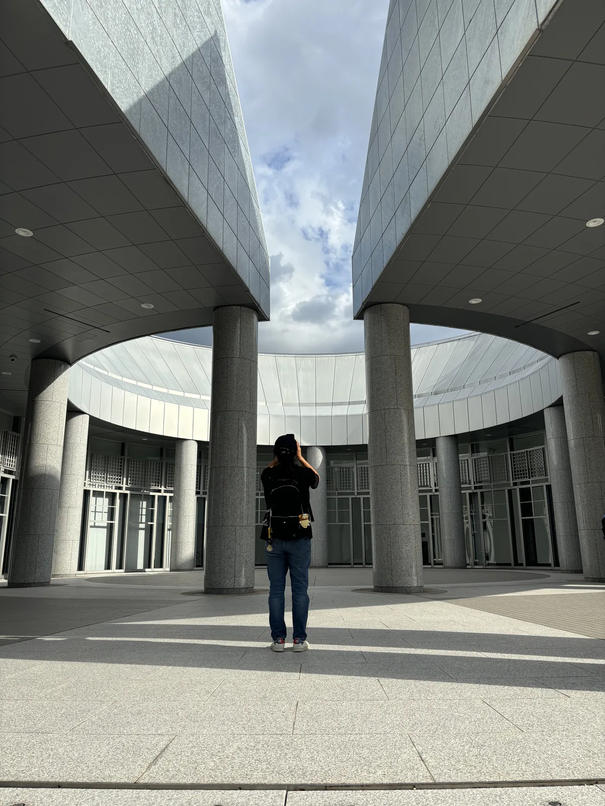 自 approach plaza 的入口望向廣場，兩根花崗岩圓柱之間，兩座切妻屋頂的山牆在上方相對，中央是被一圈弧形屋簷與圓柱圍住的圓形廣場，一位參觀者站在廣場中央拍照