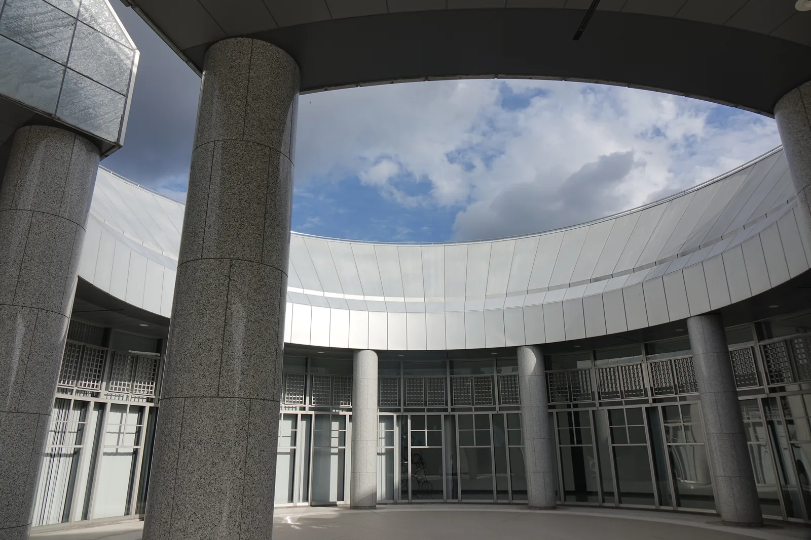 自 approach plaza 內仰望，前景是兩根粗大的花崗岩圓柱，後方是一圈銀色的弧形屋簷與圓柱列，屋簷在上方圍出一個橢圓形的天空，下方是入口的玻璃面