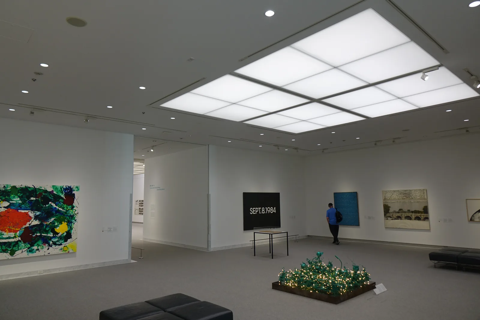 collection 展示室，天花板中央是一組以格子分割的發光天井，白色的牆上掛著繪畫與一件寫著日期的黑色作品，地面是灰色地毯，中央地上是一件小型裝置作品