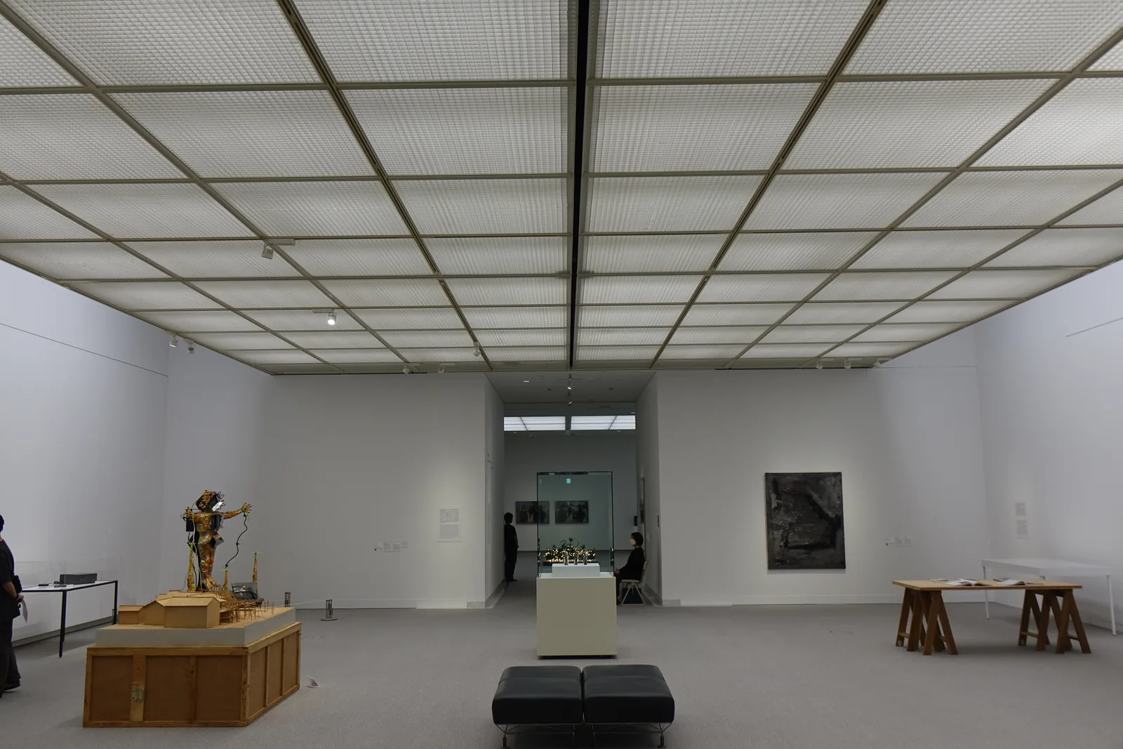 另一間展示室，整面天花板是格子狀的光天井，白色的牆與灰色地毯，中央是一件木造的作品，盡端開著一個通往下一室的門口