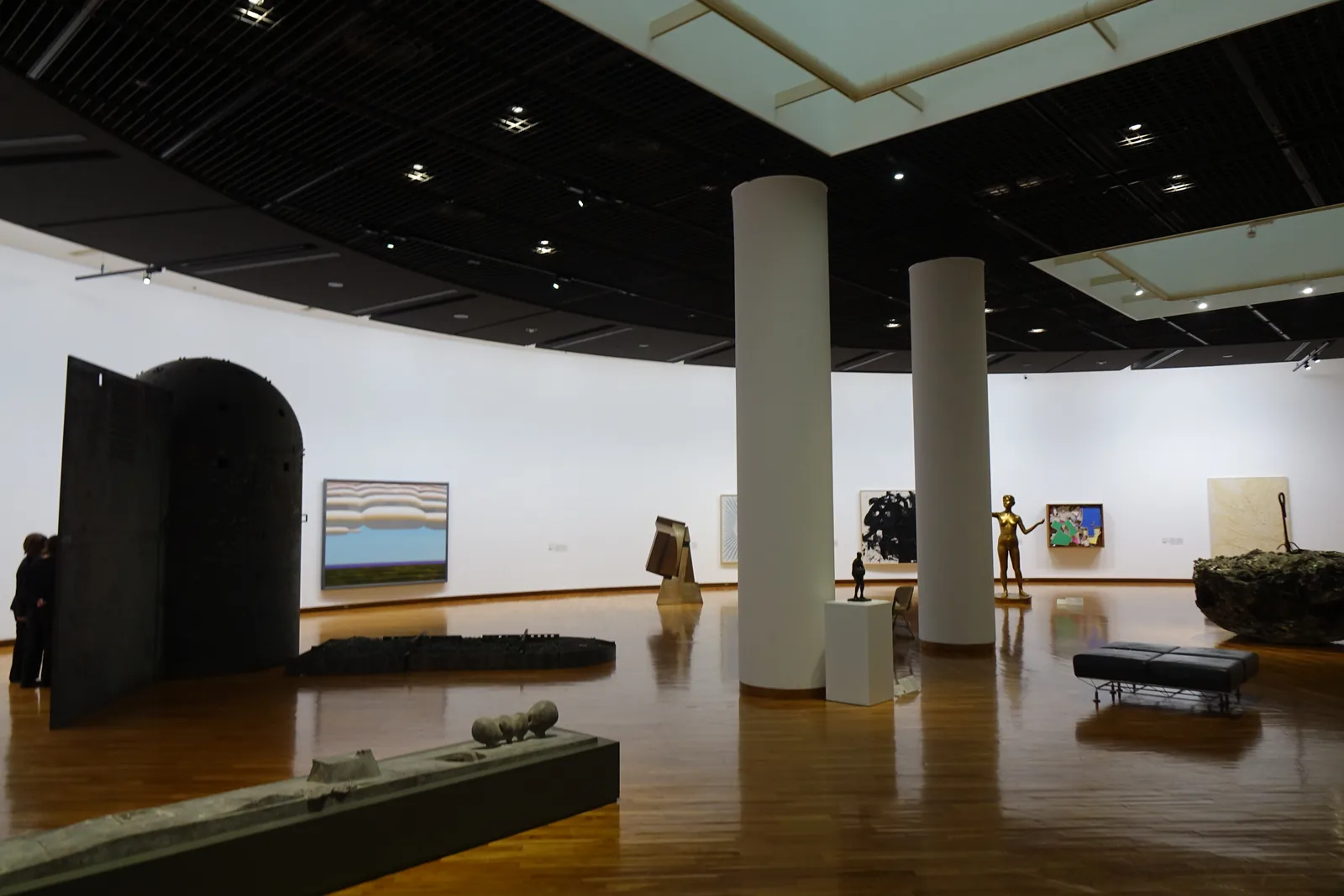 一間弧形的大展示室，地面是光亮的木地板，天花板是黑色並嵌著白色的光天井，室內立著兩根白色圓柱，左側是一件大型的黑色拱形雕刻，牆上掛著繪畫