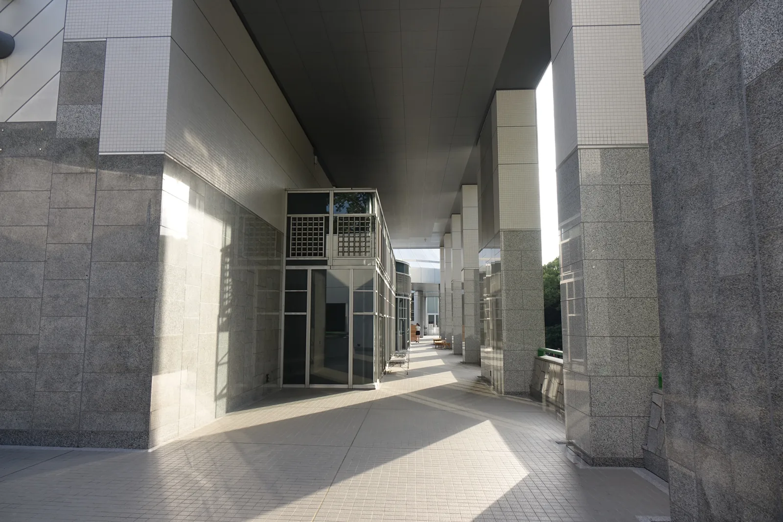 沿建築外側的柱廊，左側是以石材與磁磚包覆的方柱，右側是石材的牆面，天花板是銀色的金屬板，柱廊盡端是明亮的出口