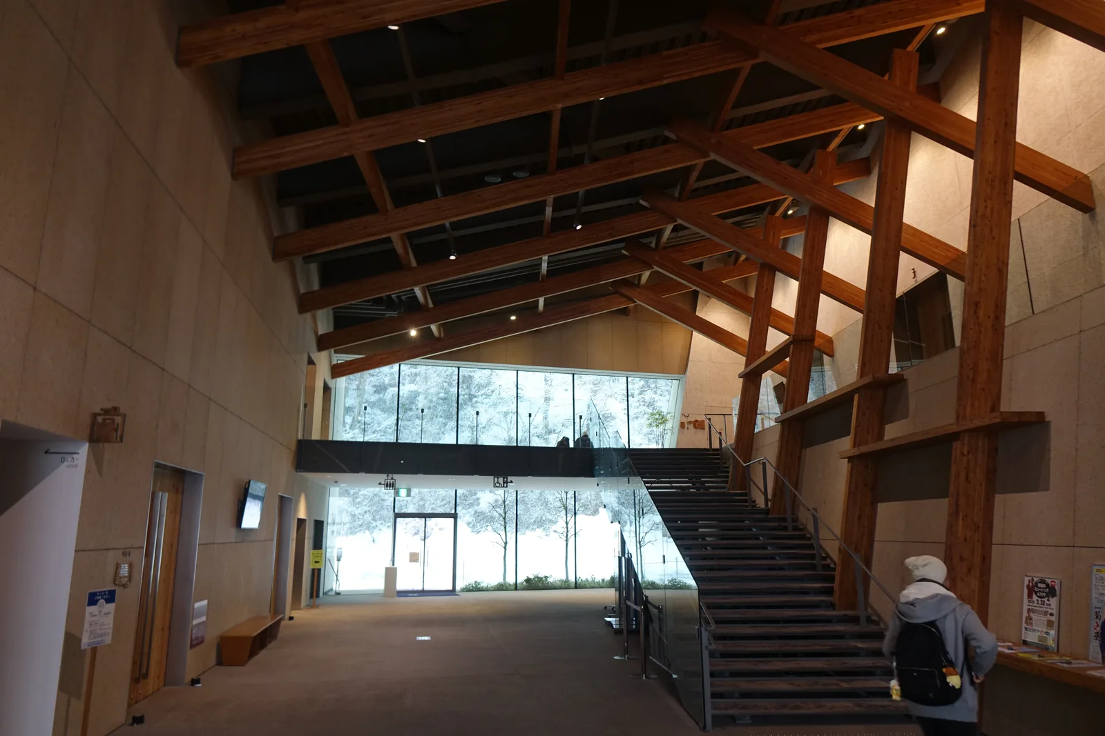 室內通道的全景，兩側牆面斜向傾倒，牆前是交錯的木構架，盡頭是一座通往二樓的鋼樓梯與一整面玻璃，玻璃外是覆雪的樹林，一位參觀者背著背包走向樓梯