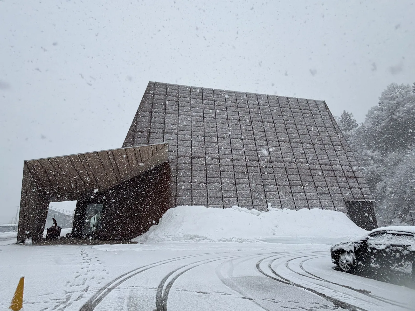 降雪中的建築全景，一個以耐候鋼覆蓋、向上斜收的大型量體，前方是一個較低的落葉松與耐候鋼量體，量體上有幾處開口，量體腳下堆著鏟起的積雪，前景是積雪的停車場與輪胎痕
