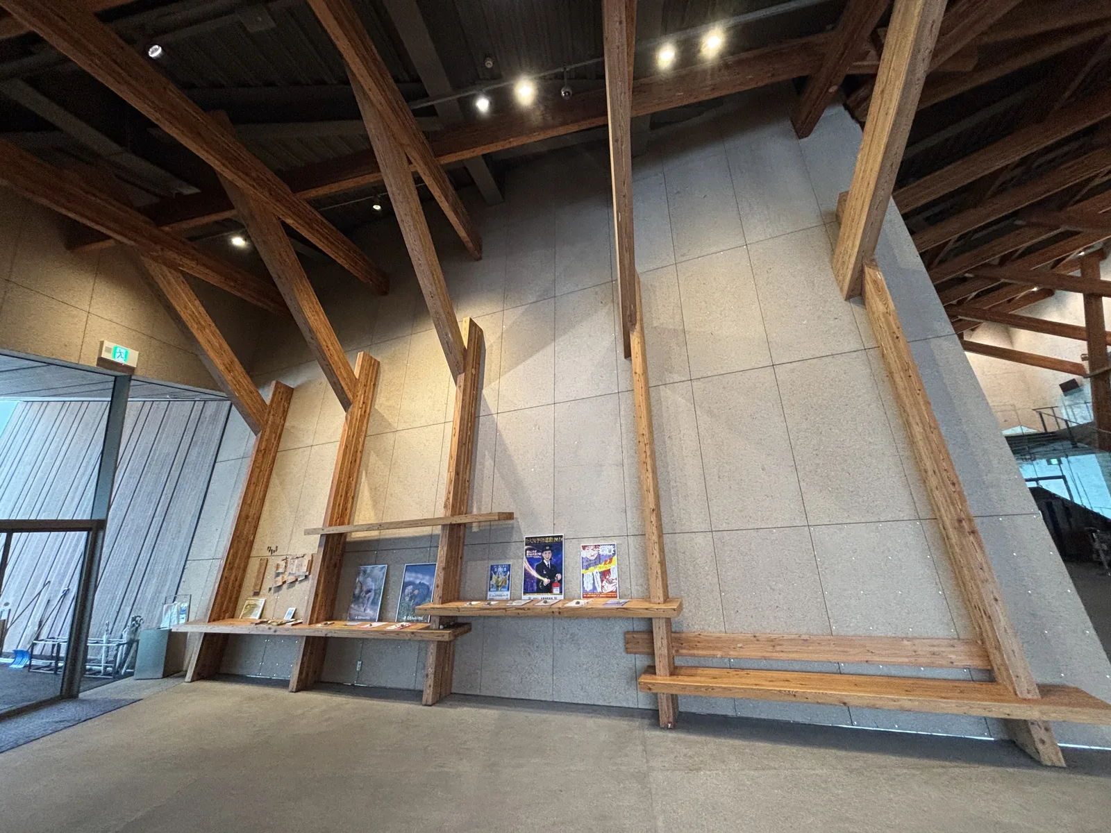 中道的一面牆，灰色的粗面板材向內傾斜，牆前是斜向的木構架，構架之間安裝著木製層架，層架上放著傳單與節目單，上方是深色的鋼梁與照明