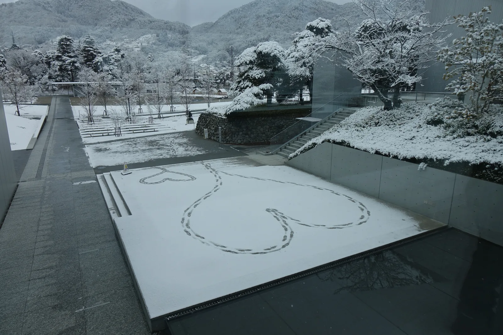 自連絡橋俯瞰的水邊露台，平台覆著一層薄雪，雪上有人踩出的心形足跡，平台以幾段階梯向後方退去，右側是一道石垣與通往上方的石階，後方是公園、覆雪的樹木與山