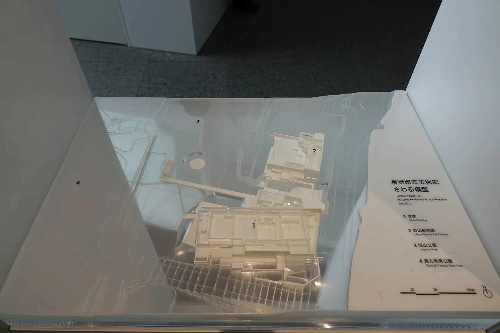 館內展示的建築模型，白色的模型呈現本館的量體、連絡橋與階梯狀的水邊露台，放在一個斜向的白色台座上，台座一側貼著寫有模型說明的標籤