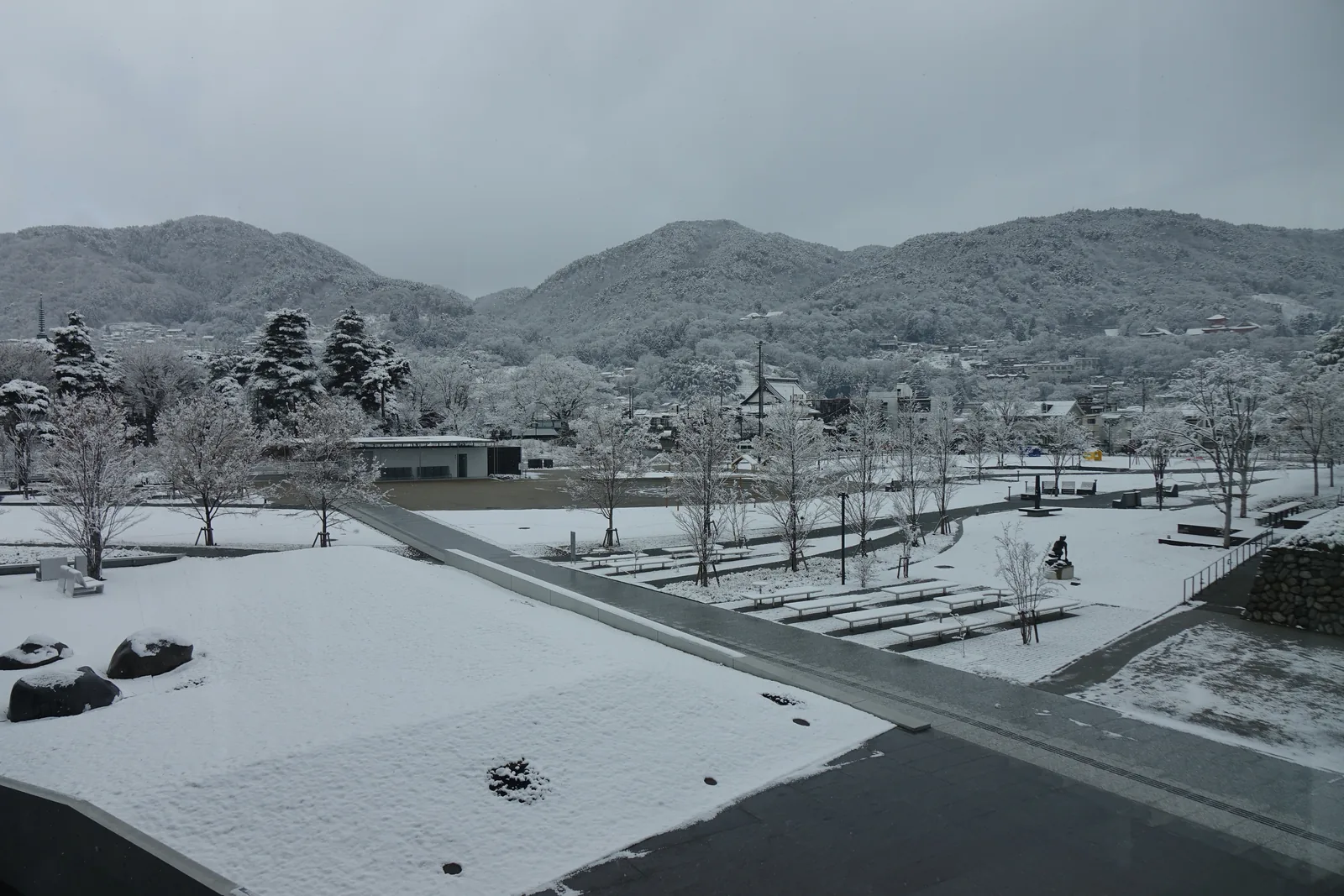 自館內高處望向城山公園，前景是覆雪的屋頂平台與幾塊景石，中景是一條直線的步道與整排的長椅、雕塑和樹木，遠景是覆雪的山與山腳下的街區