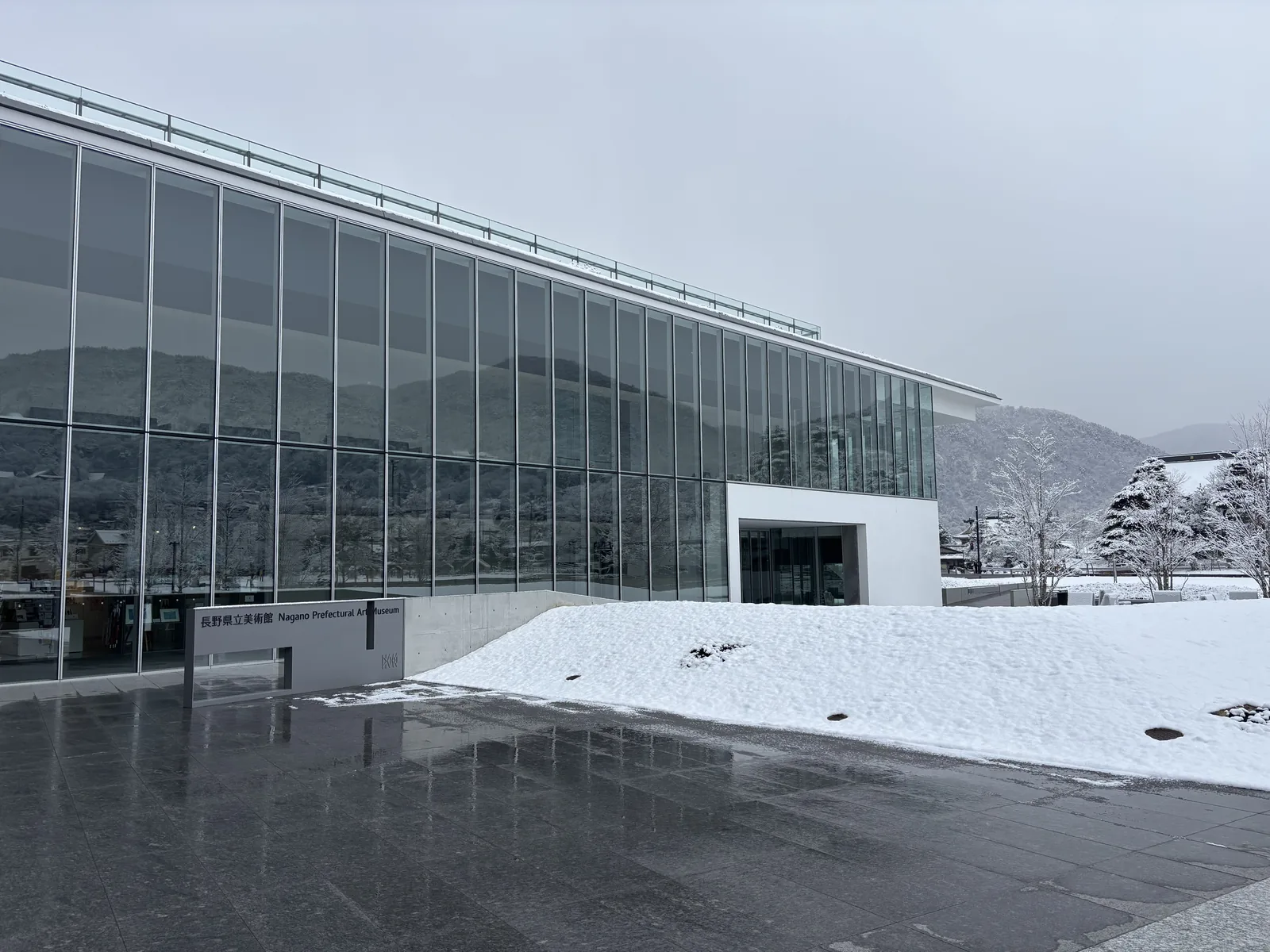 美術館的入口立面，一道長而低的玻璃 curtain wall 反射著天空與山，右側是一個白色的方形量體與入口，前方是濕潤的石材廣場與一片覆雪的草坡，左側立著館名的石碑