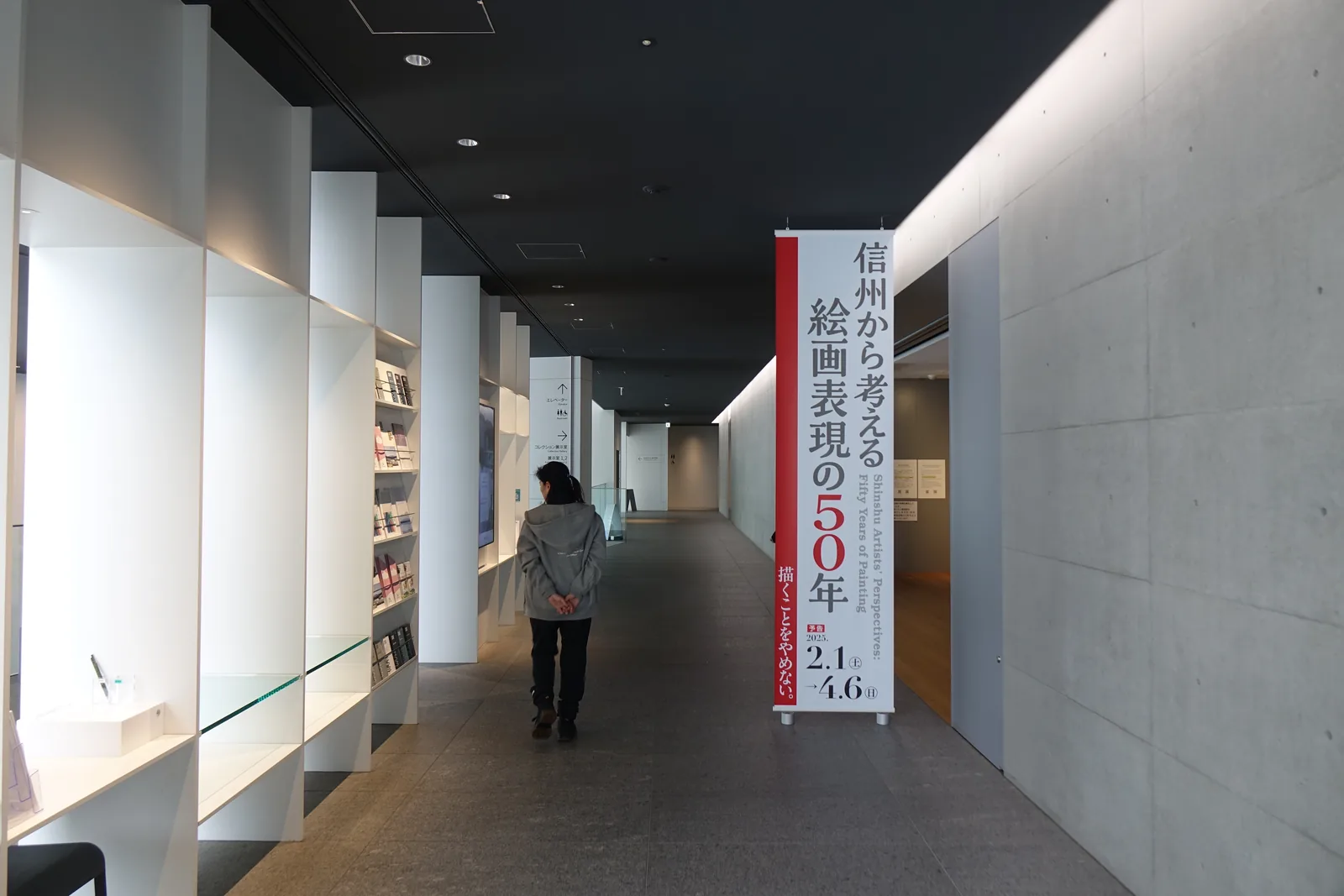 一條通往展示室的走廊，左側是一排白色的展示櫃，嵌著介紹資料，中央掛著一幅寫有展覽名稱的紅白直幅，右側是一整面清水混凝土牆，一位參觀者沿走廊走向遠處