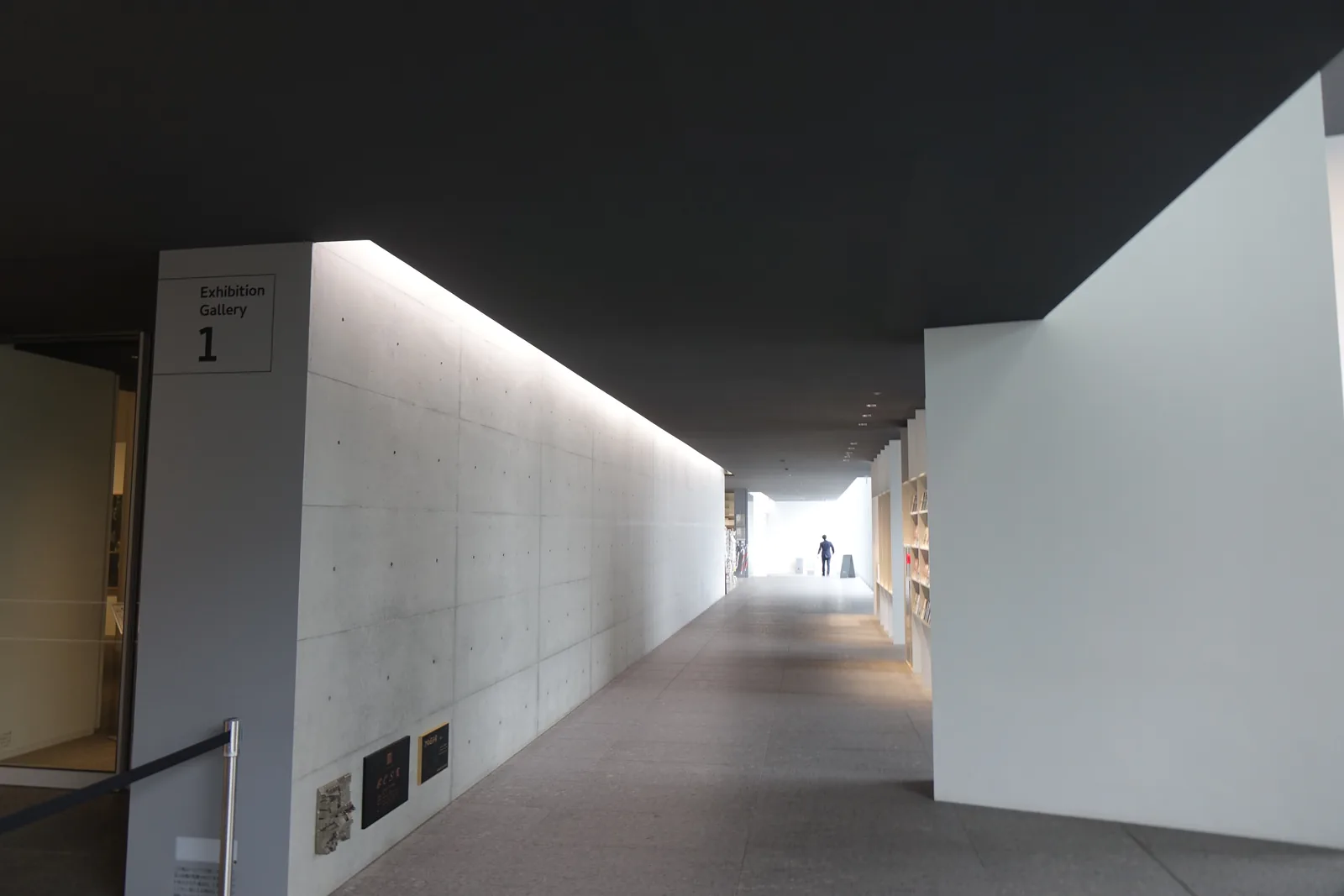 一條通往展示室一的走廊，左側是一道白色的牆與展示室一的標示，右側是一道白色的牆端，深色的天花在走廊上方形成一道帶狀，走廊盡頭是一個明亮的開口與一位行人