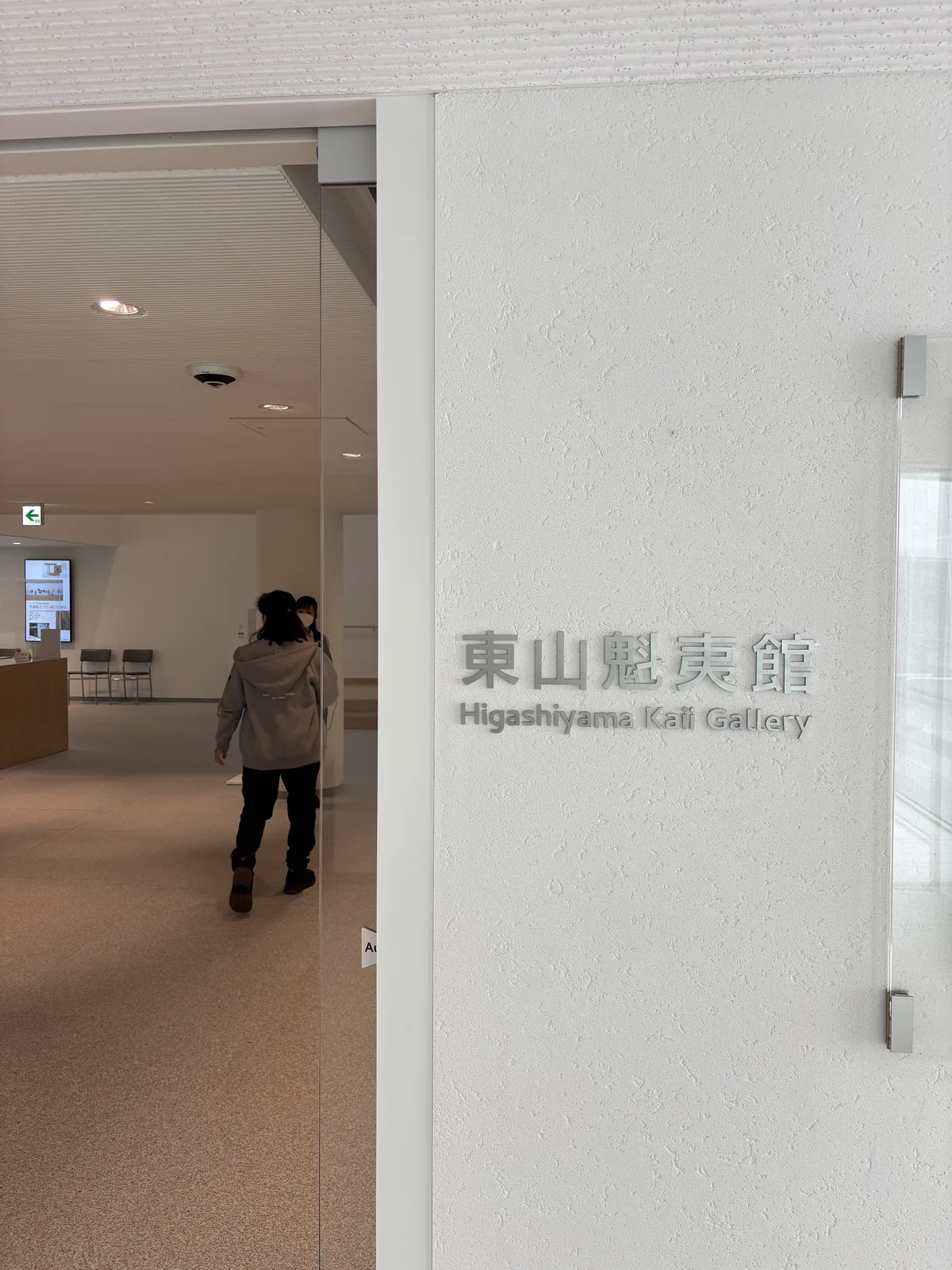 東山魁夷館入口的玻璃門，門上以灰色字印著館名與英文館名，門內是淺色的木地板與一條明亮的走廊，一位參觀者走進門內