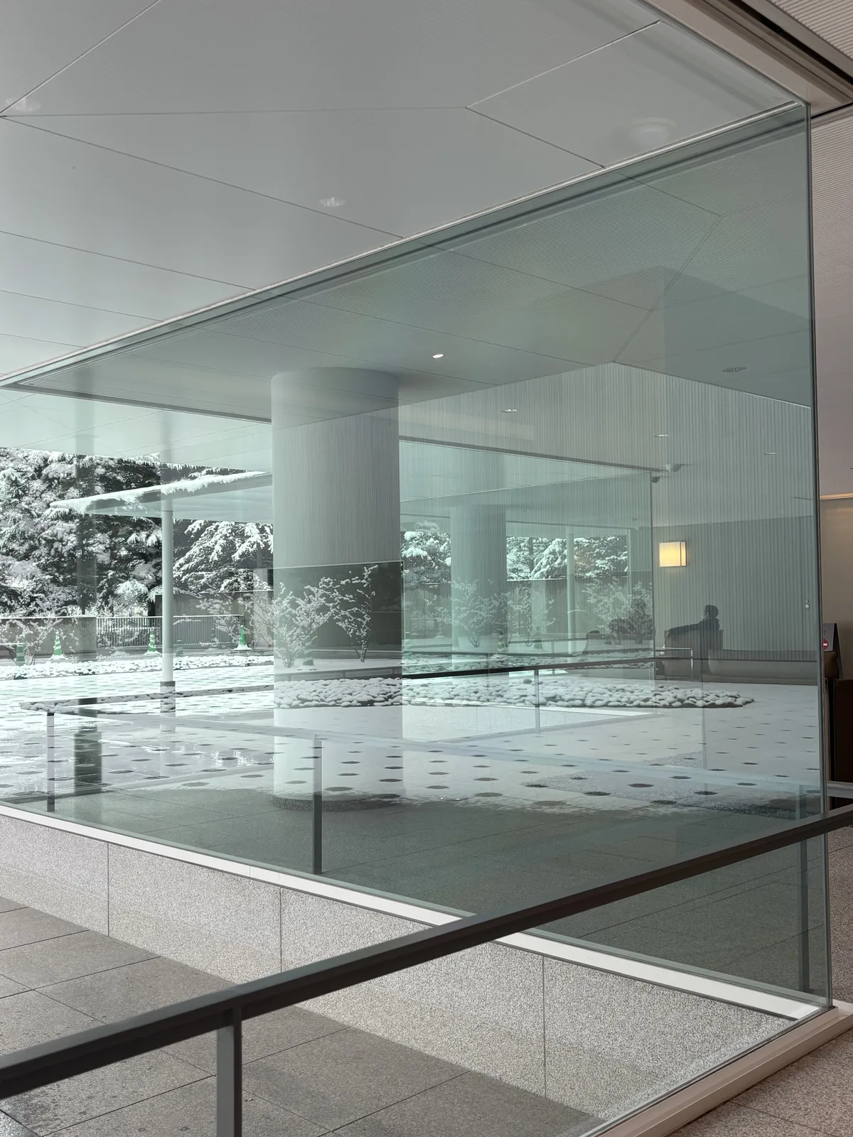 東山魁夷館的 lounge，一面玻璃牆面對中庭，玻璃外是一根白色的圓柱與覆雪的中庭，玻璃內是淺色的石材地面與一道玻璃扶手，遠處坐著一位參觀者