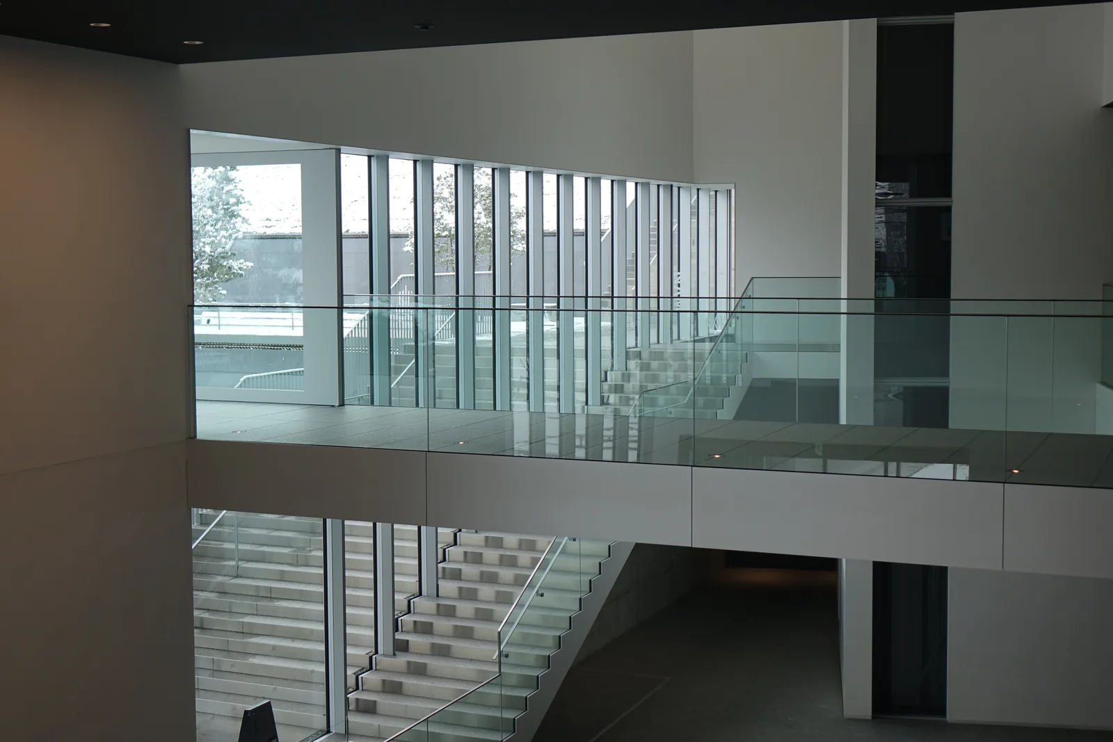 自本館內部望向連絡橋，前景是一道玻璃扶手，中景是連絡橋的玻璃通道與下方的階梯，玻璃外是覆雪的樹木，上方是深色的天花與一排細長的直條