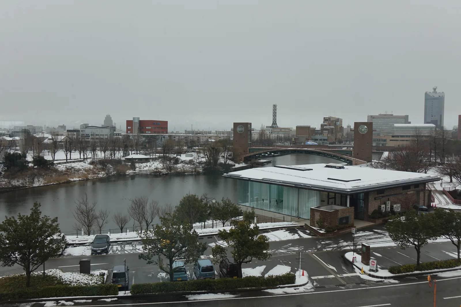 自三樓的窗戶俯瞰富岩運河環水公園，前景是一條道路與停車場，中景是運河的水面與水邊一棟平屋頂的建築，遠景是富山的市街、一座高樓與幾座塔，覆雪的屋頂散落在各處
