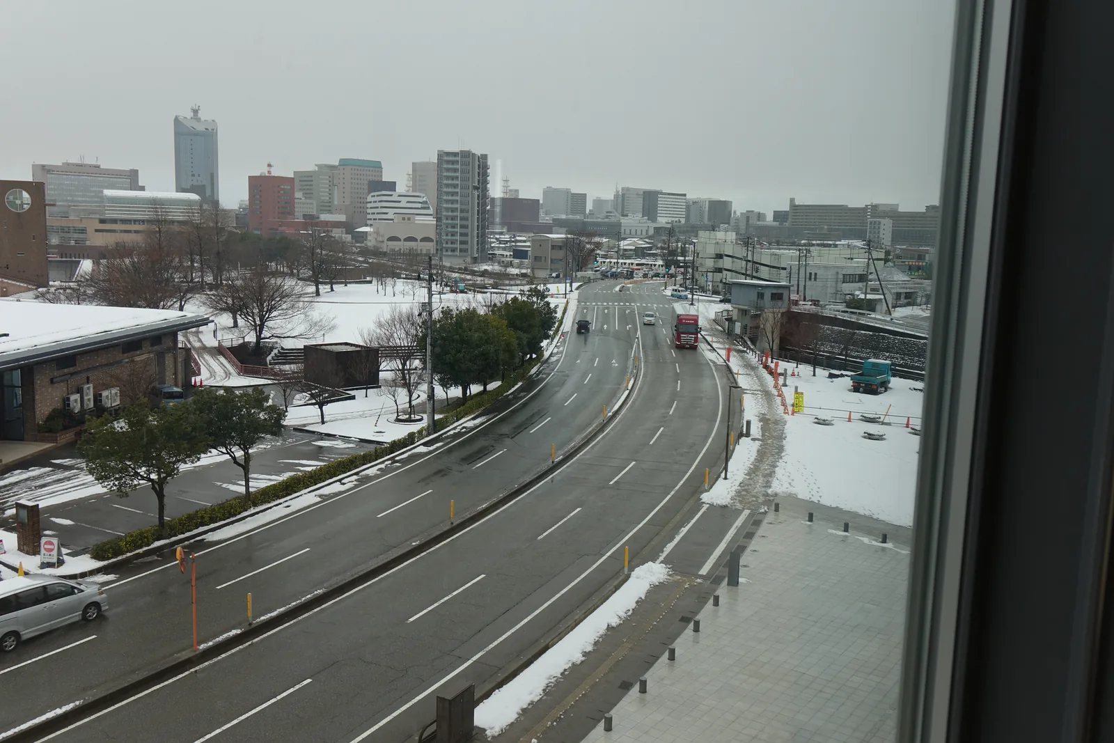 自三樓的窗戶望向南方的道路，一條寬闊的道路自前景向遠處彎曲延伸，兩側是覆雪的人行道與樹木，遠處是富山的市街與一座高樓，右側是窗框