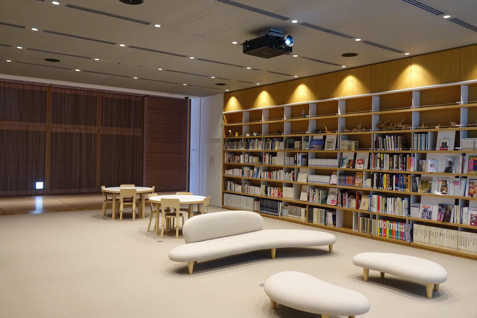 三樓的 hall，一整面牆是排滿書籍的木製書架，書架上方有一排間接照明，中央是幾張圓桌與木椅，前景是幾件白色的曲線長椅，天花上懸著一台投影機