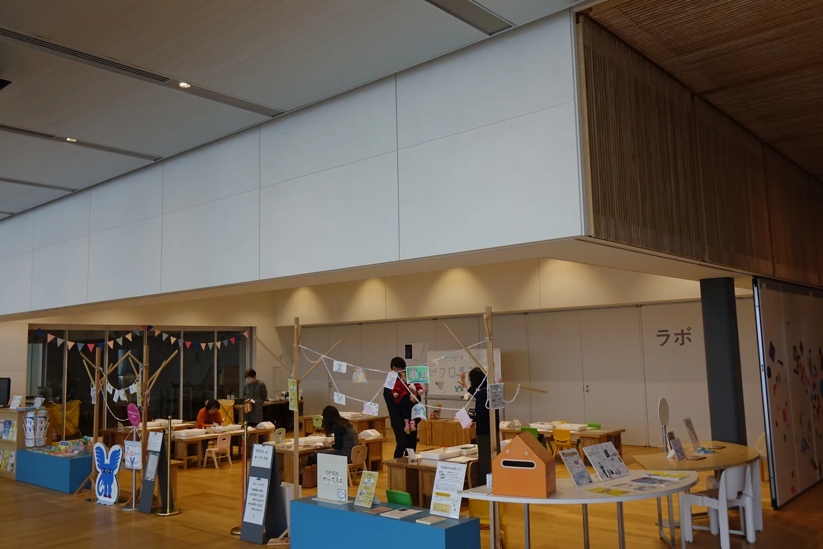 三樓的 atelier，玻璃隔間內是幾張工作桌與放滿材料的層架，幾位大人與孩子正在桌邊製作，上方是白色的牆面與木條的端部，入口處立著一塊看板